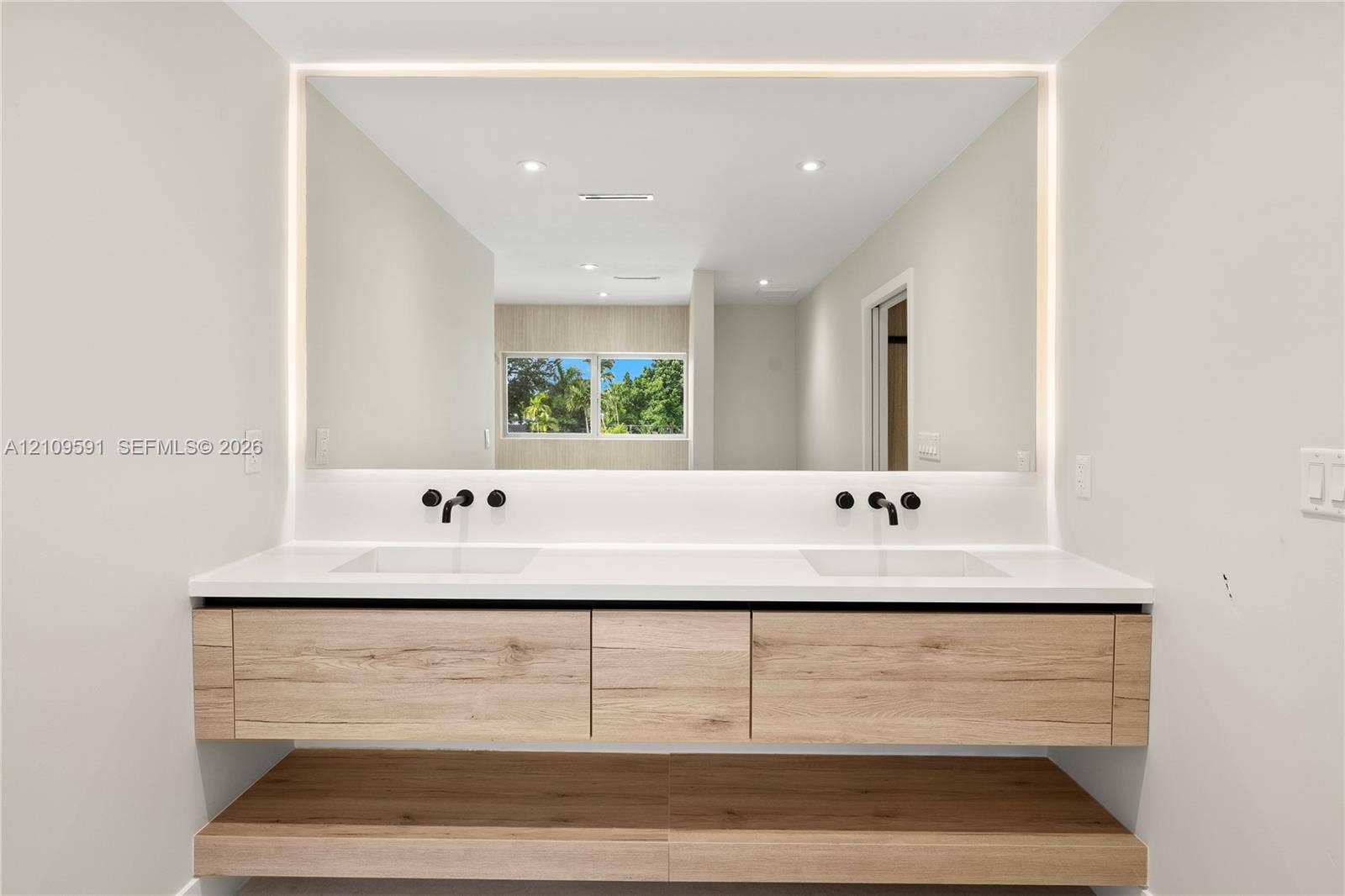 864 NE 91st Terrace, Miami Shores, FL 33138 Floating wood-finish vanity with dual integrated sinks, white quartz countertop, matte black wall-mounted faucets, and large LED-backlit mirror