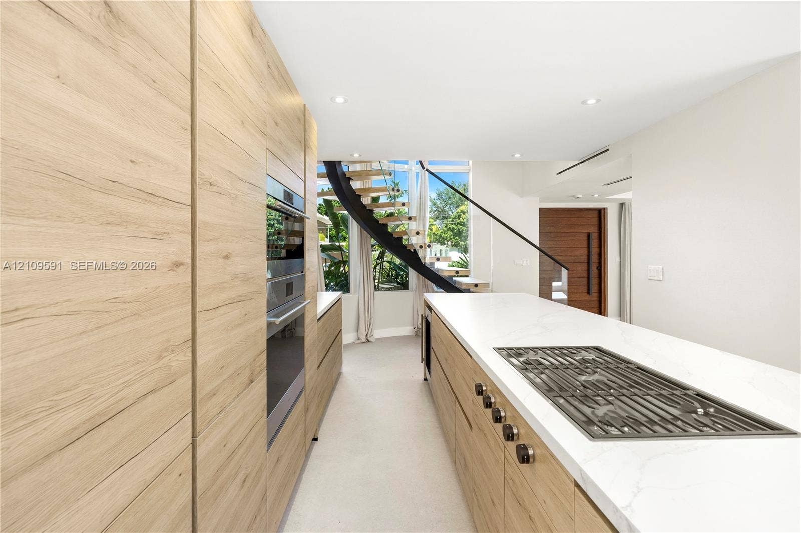 864 NE 91st Terrace, Miami Shores, FL 33138 Kitchen featuring full-height wood-finish cabinetry, integrated wall oven, large island with quartz-style countertop, inset gas cooktop, and a curved staircase with floating wood treads