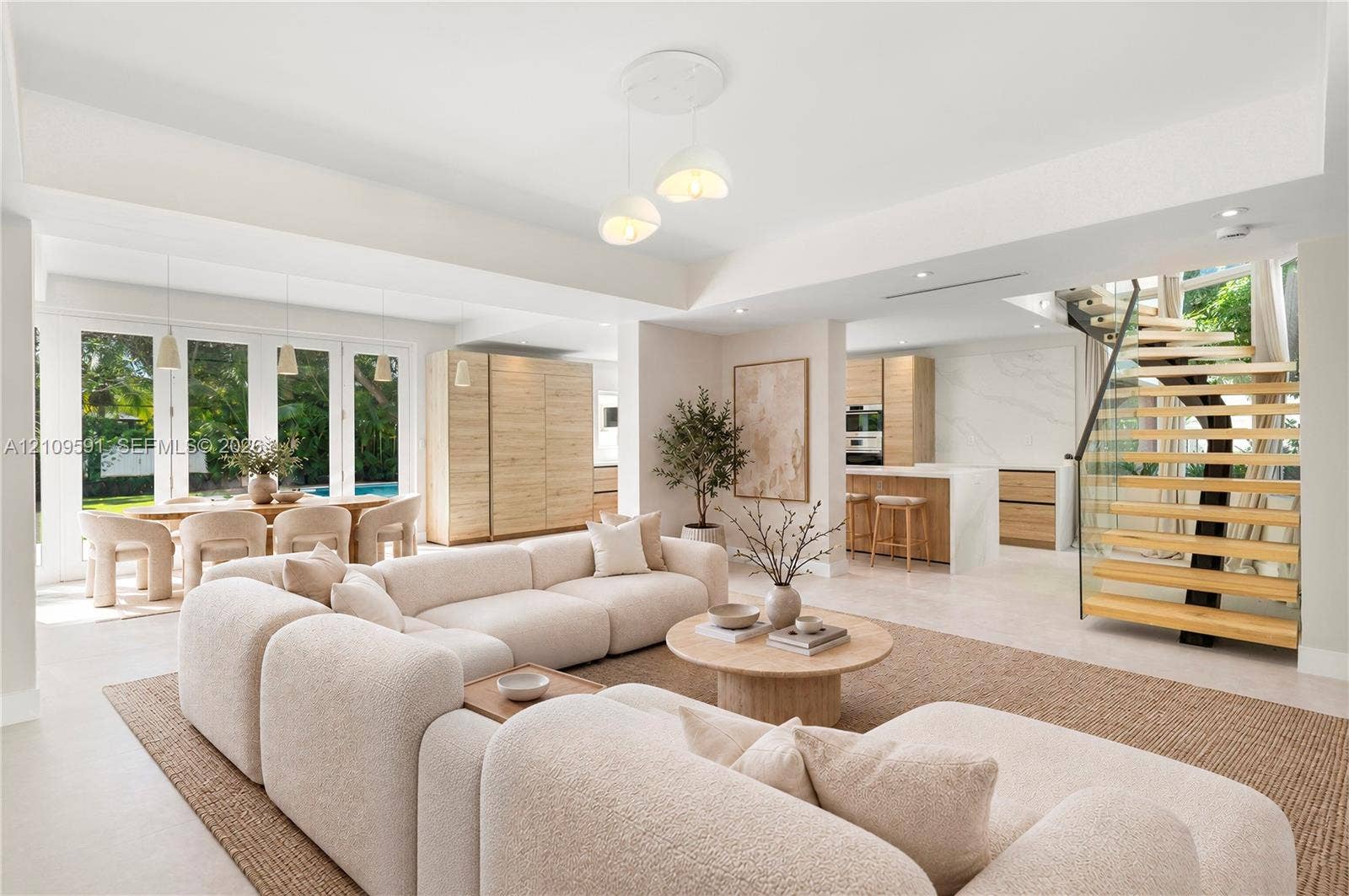 864 NE 91st Terrace, Miami Shores, FL 33138 Virtually Staged: Open-concept living space featuring high tray ceilings, wood-finish cabinetry, a modern staircase with glass railings and wood treads, large-format tile flooring, and multiple glass-paneled exterior doors