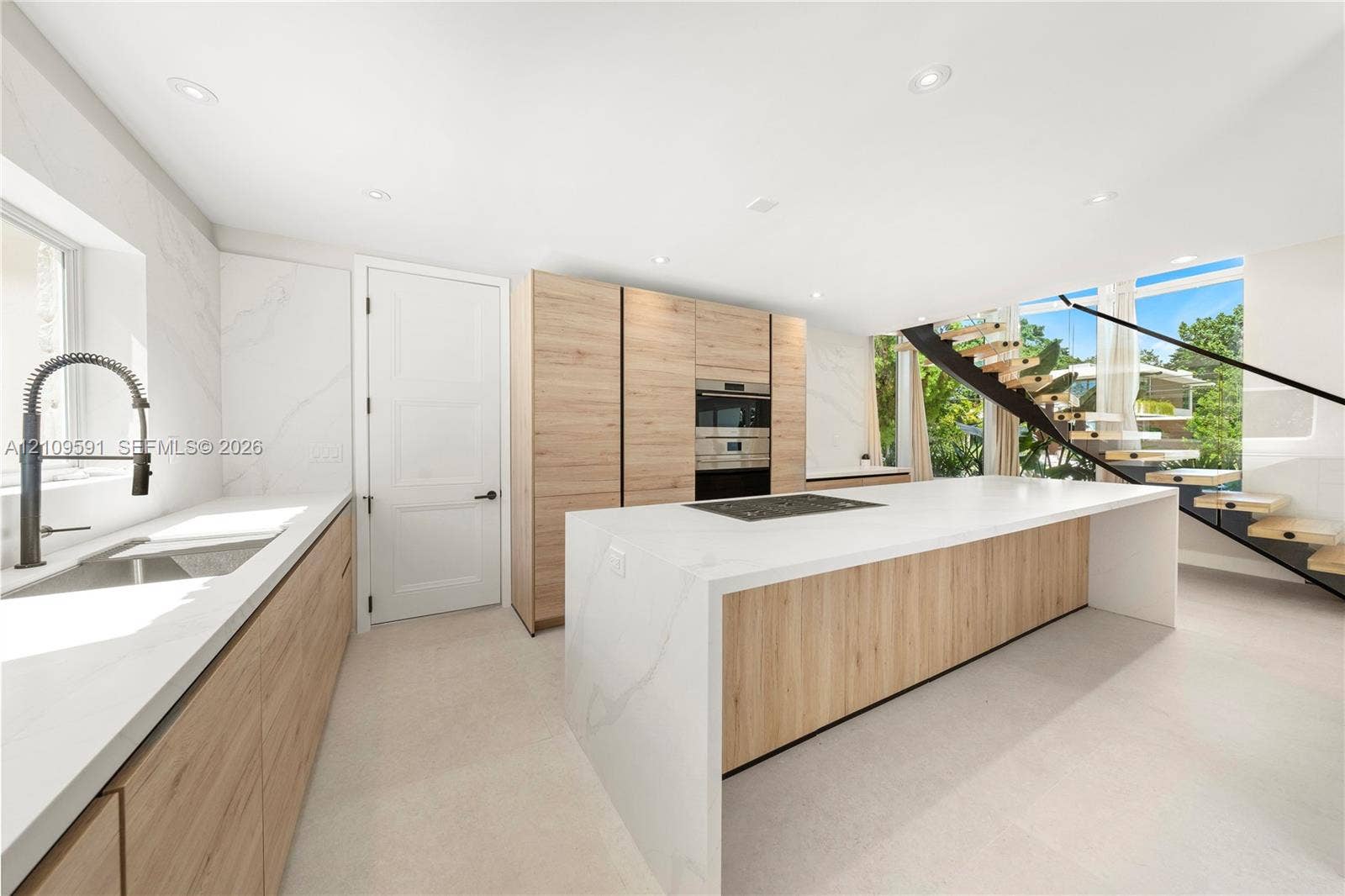 864 NE 91st Terrace, Miami Shores, FL 33138 Modern kitchen featuring a waterfall-edge island, light wood-finish cabinetry, stone-style countertops, recessed lighting, and a floating staircase with black metal railings