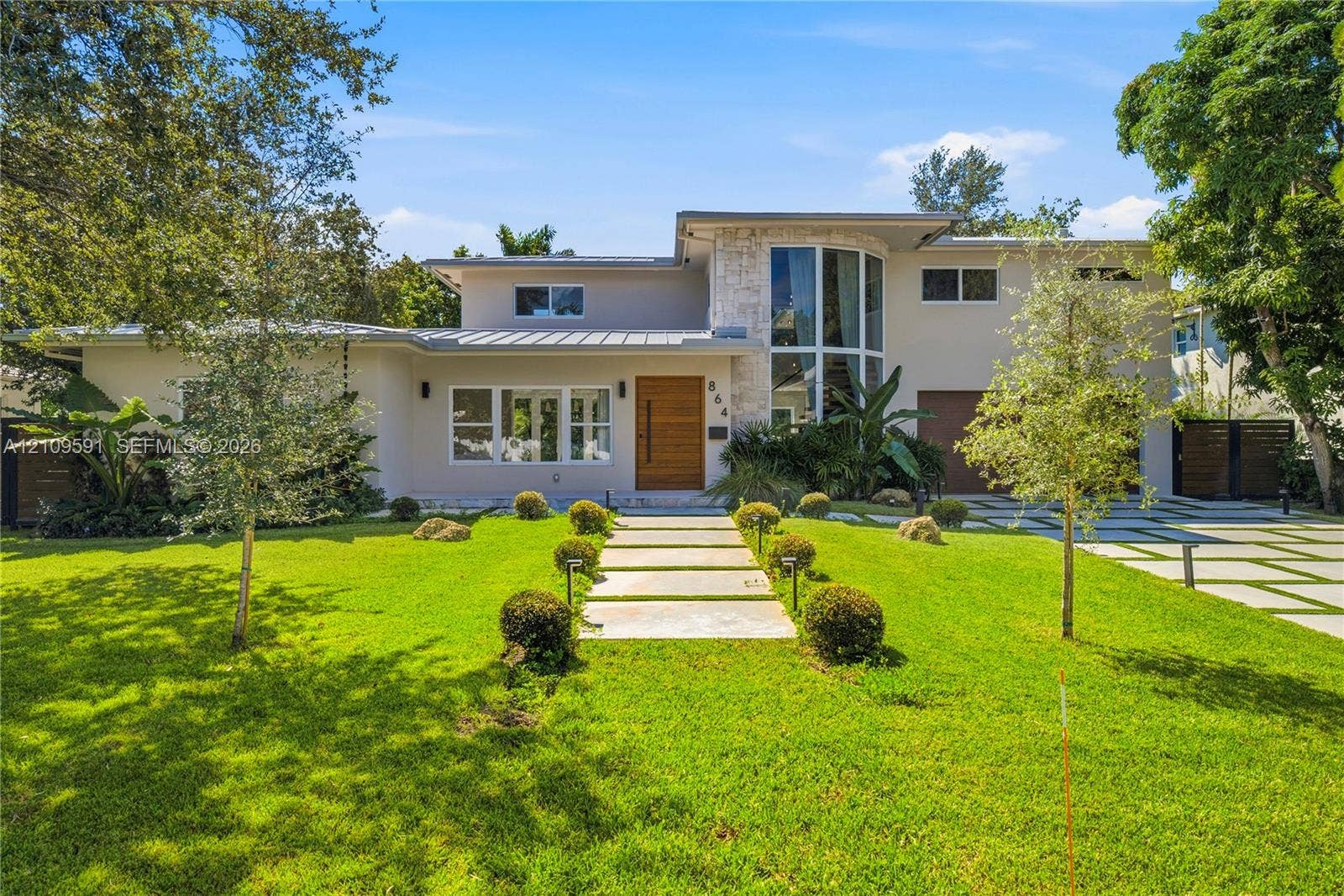 864 NE 91st Terrace, Miami Shores, FL 33138 Modern exterior featuring a metal roof, stone facade accents, expansive floor-to-ceiling windows, a wood-finish front door, and a geometric concrete paver driveway