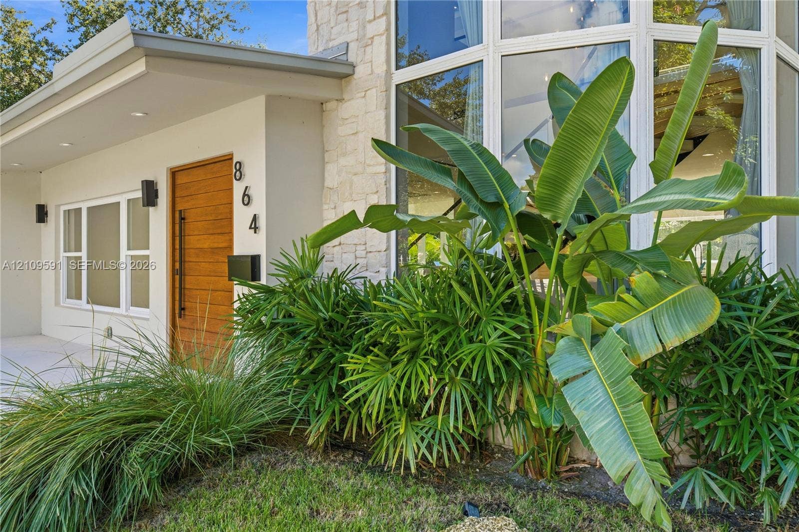 864 NE 91st Terrace, Miami Shores, FL 33138 Modern facade featuring a natural stone accent wall, expansive multi-pane windows, and a horizontal wood-grain door with a vertical matte black pull handle