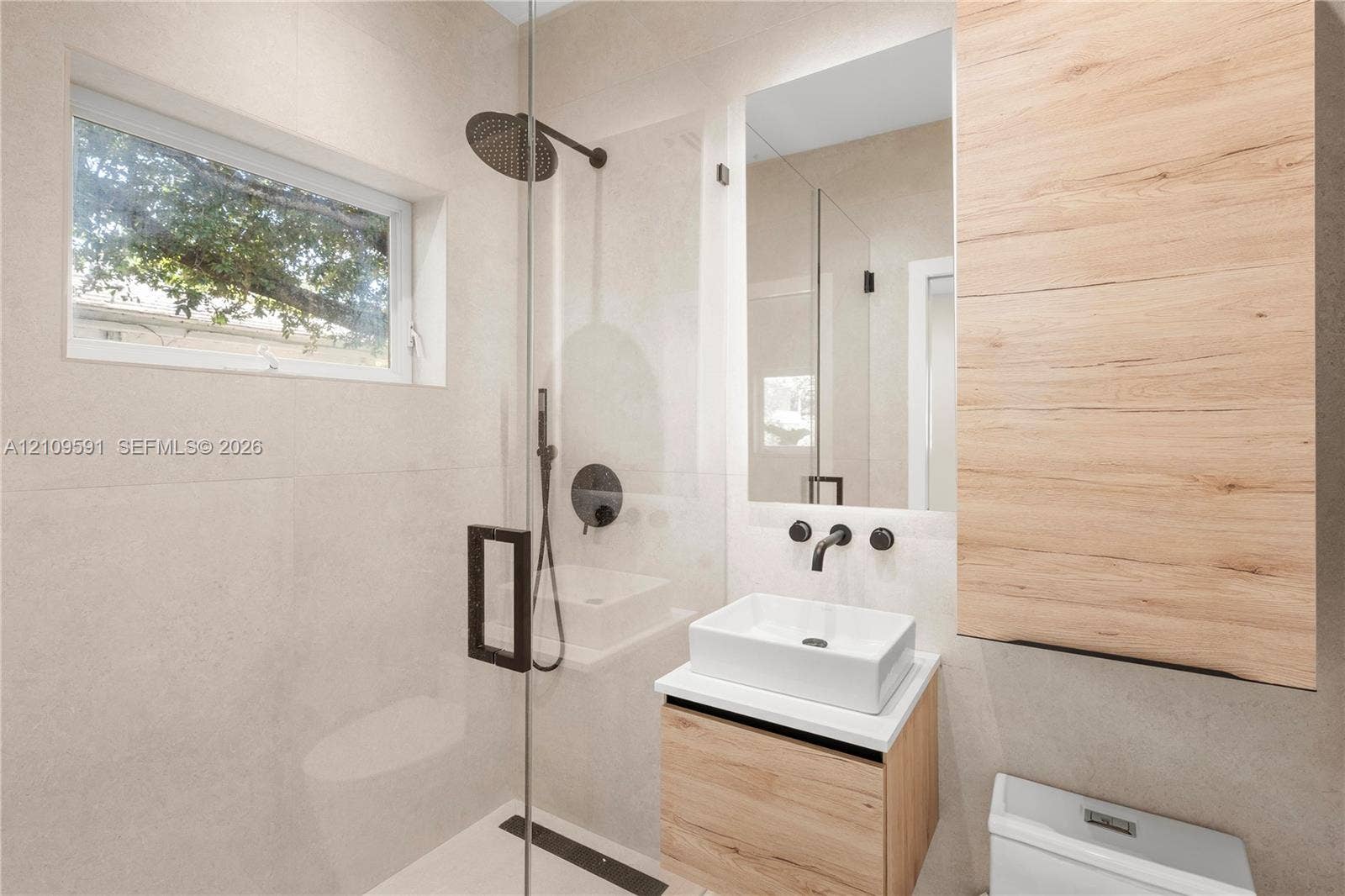 864 NE 91st Terrace, Miami Shores, FL 33138 Modern bathroom featuring large-format wall tiling, a floating wood-finish vanity with a rectangular vessel sink, matte black hardware, and a glass-enclosed shower with a rainfall showerhead