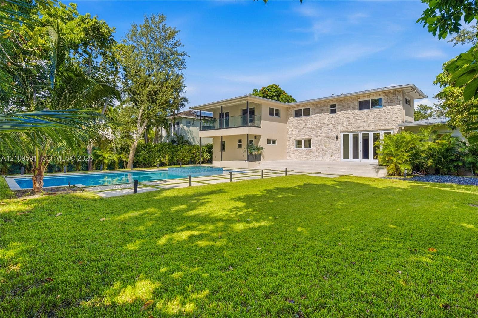 864 NE 91st Terrace, Miami Shores, FL 33138 Exterior facade featuring stone veneer cladding, swimming pool with integrated spa, glass-panel balcony railing, and manicured lawn