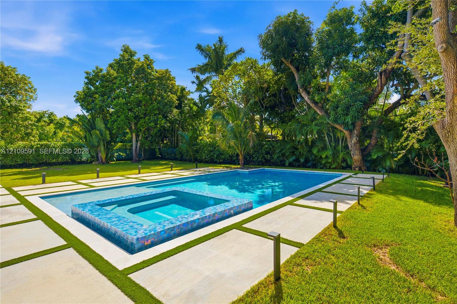 864 NE 91st Terrace, Miami Shores, FL 33138 In-ground swimming pool featuring a tiled spillover spa, large-format concrete paver decking, manicured lawn, and mature perimeter landscaping