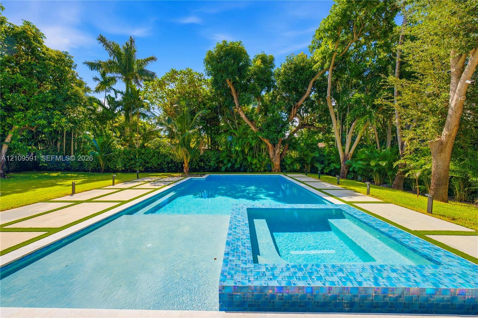 864 NE 91st Terrace, Miami Shores, FL 33138 In-ground swimming pool with an integrated spa, mosaic tile finish, concrete paver decking, and mature landscaping