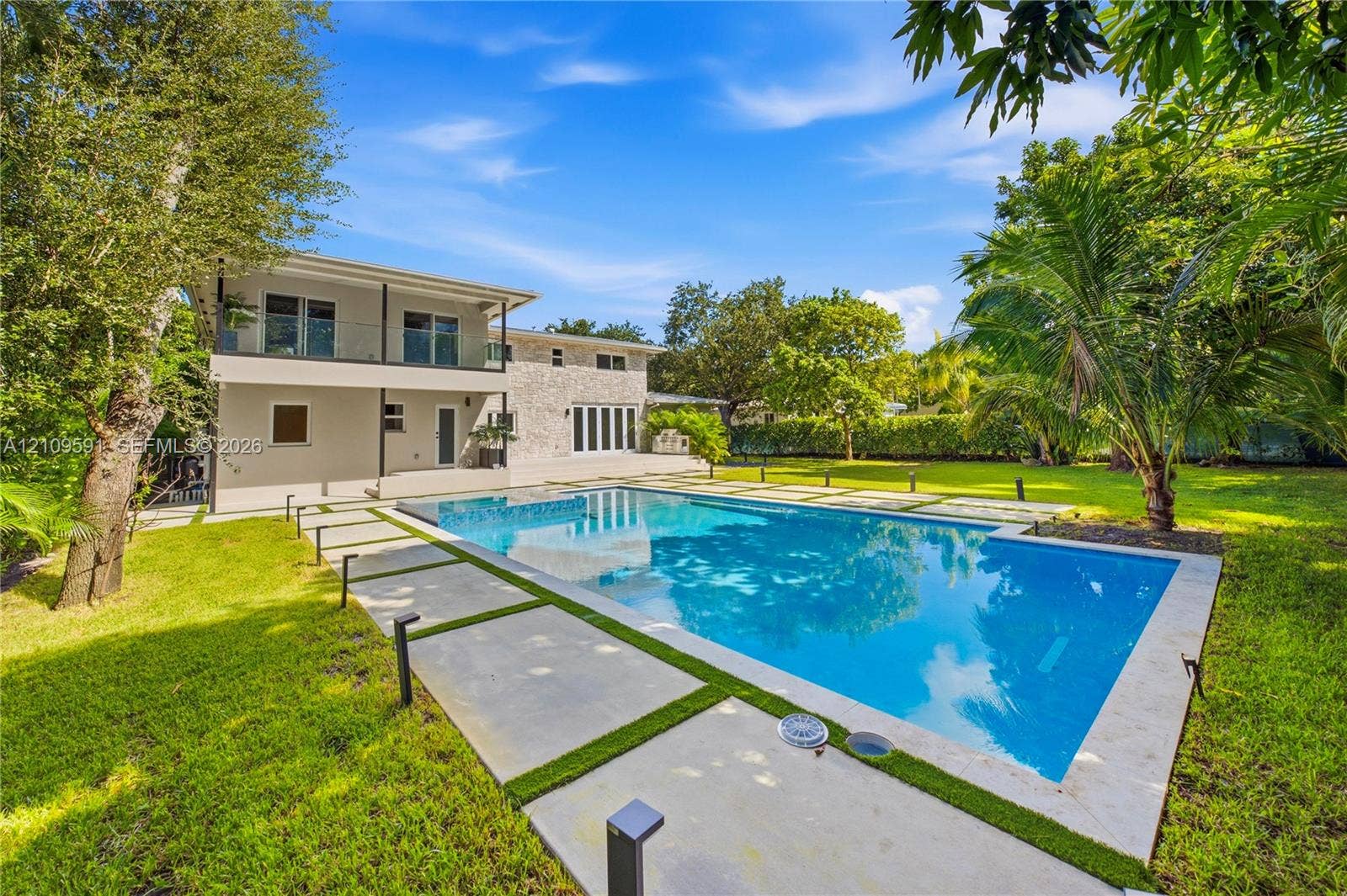 864 NE 91st Terrace, Miami Shores, FL 33138 Backyard featuring a rectangular swimming pool, concrete paver decking, manicured lawn, stone-veneer exterior finish, and a second-level balcony with a glass railing