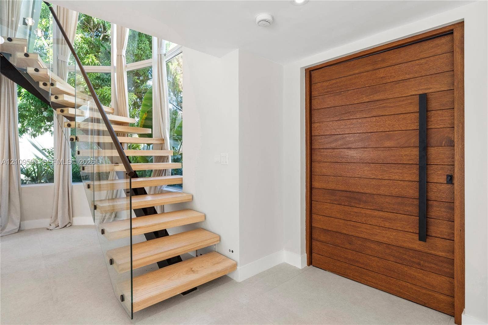 864 NE 91st Terrace, Miami Shores, FL 33138 Entryway featuring a horizontal-plank wood door with a vertical matte black handle, a floating staircase with wood treads, and a glass panel railing