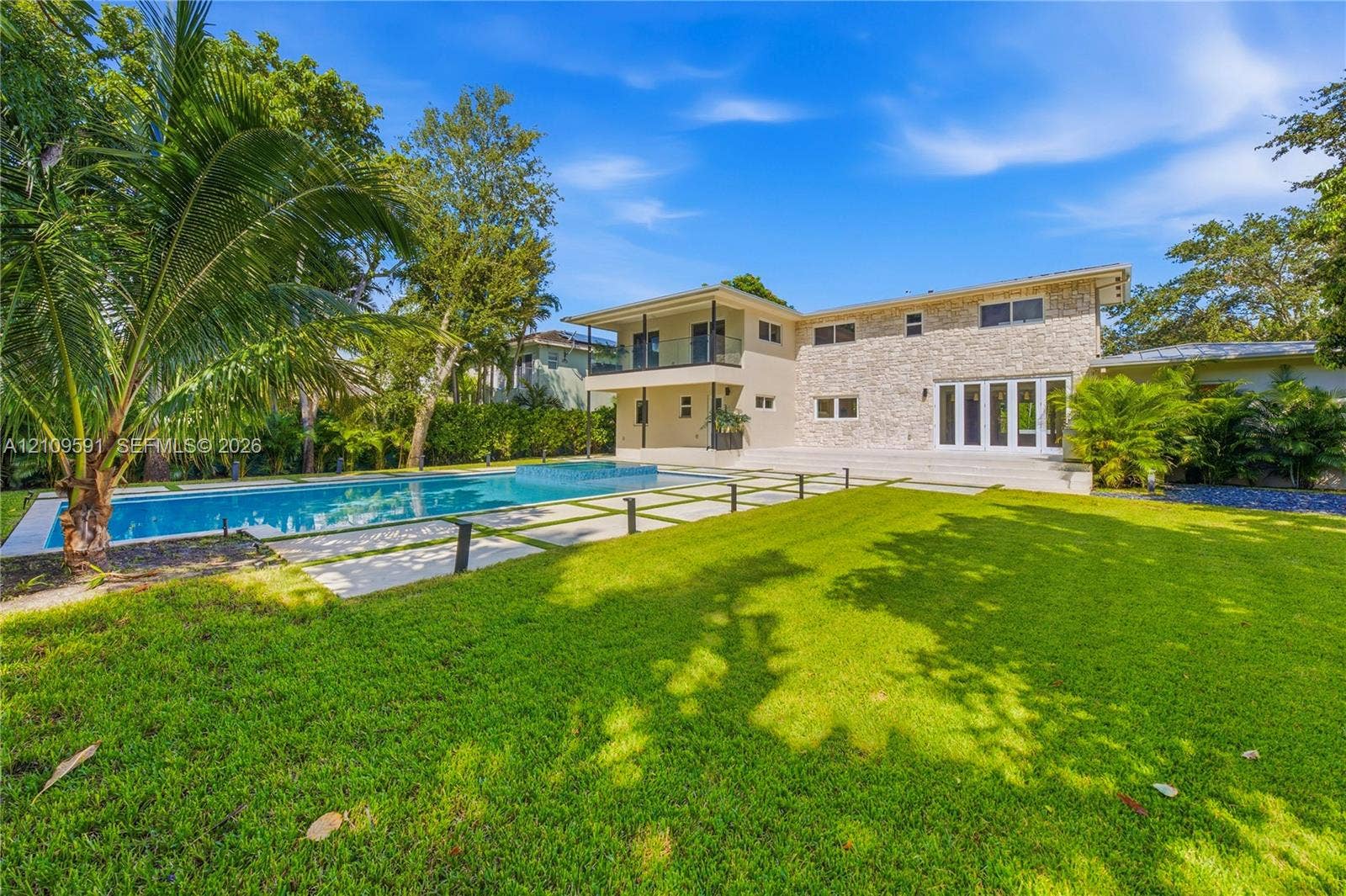 864 NE 91st Terrace, Miami Shores, FL 33138 Exterior facade featuring natural stone veneer, in-ground swimming pool with integrated spa, manicured lawn, tiered patio, and second-story balcony with glass railing
