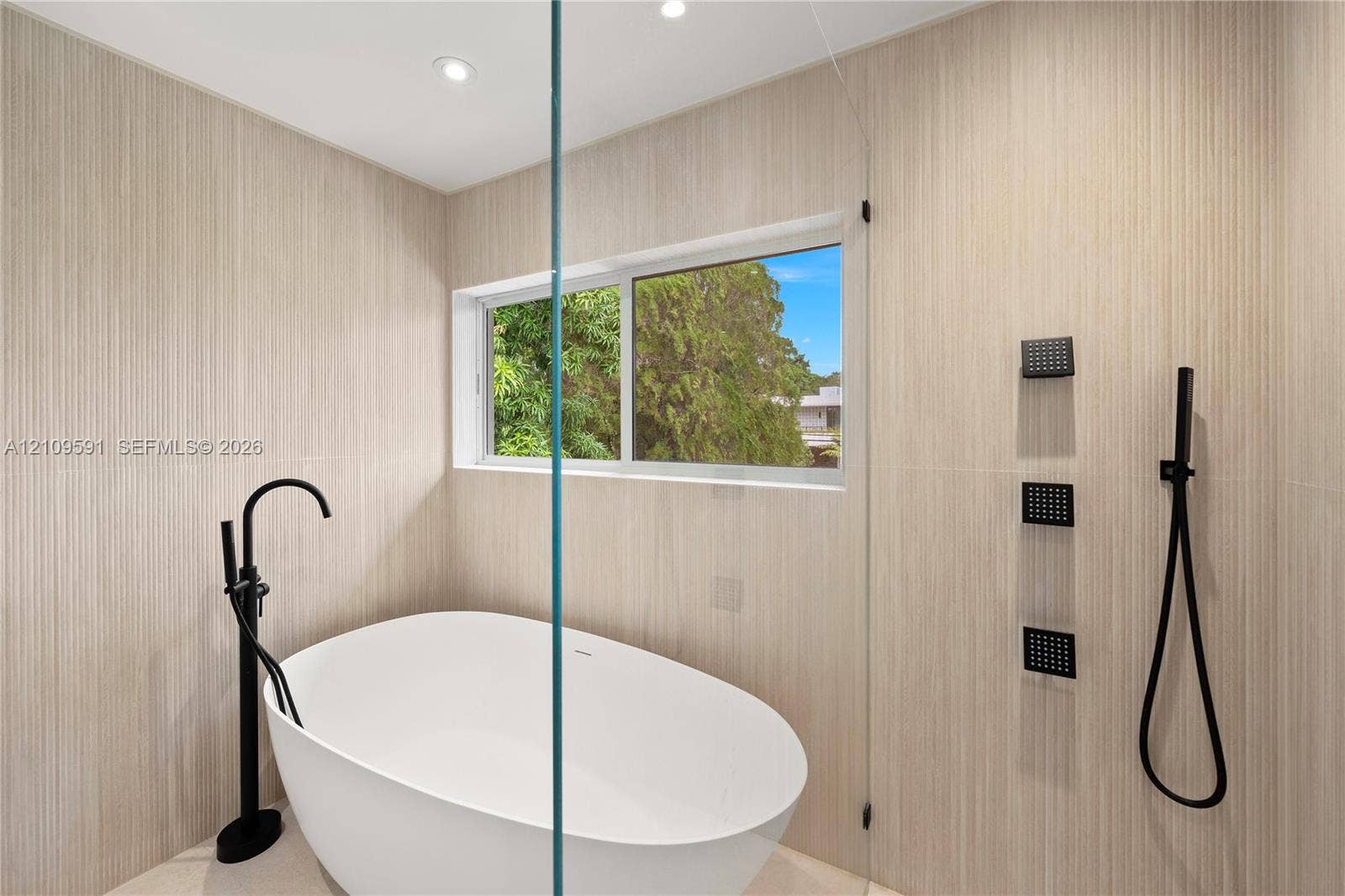 864 NE 91st Terrace, Miami Shores, FL 33138 Freestanding white soaking tub with matte black floor-mounted faucet, floor-to-ceiling textured tile wall, frameless glass shower partition, and matte black rainfall shower system with handheld sprayer