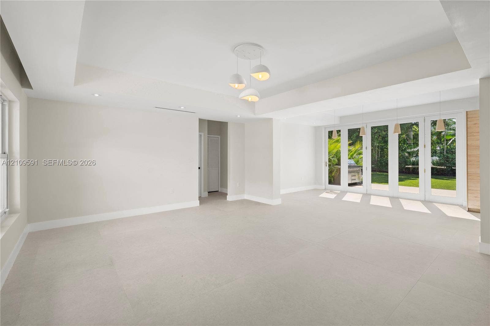864 NE 91st Terrace, Miami Shores, FL 33138 Open-concept interior featuring a recessed tray ceiling, large-format tile flooring, multi-pane glass doors, and contemporary tiered pendant lighting