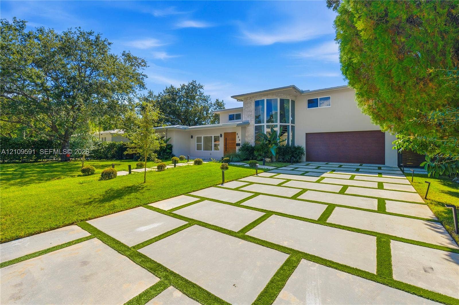 864 NE 91st Terrace, Miami Shores, FL 33138 Contemporary residence featuring a segmented concrete paver driveway, vertical multi-story glass paneling, wood-finished entrance door, and a manicured lawn