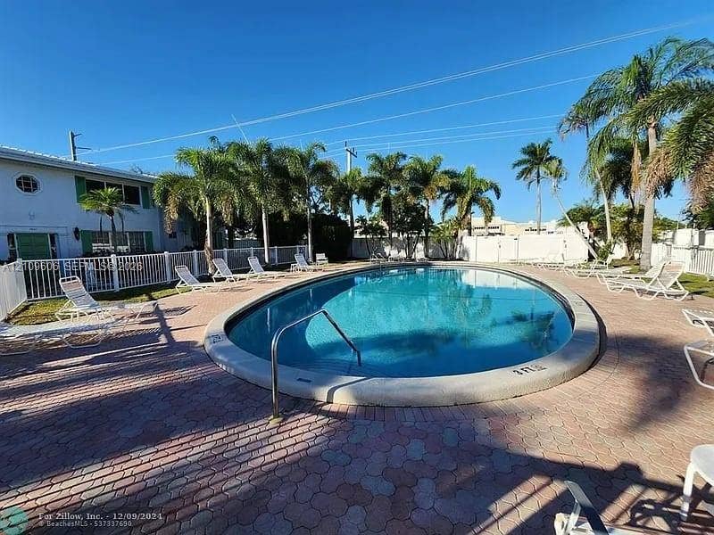 1851 NE 62nd Street, #610, Fort Lauderdale, FL 33308 In-ground swimming pool with a concrete coping and metal handrail, surrounded by a paved patio and white perimeter fencing, accented by mature palm trees