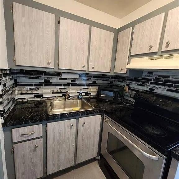 1851 NE 62nd Street, #610, Fort Lauderdale, FL 33308 Kitchen featuring light wood-finish cabinetry, dark stone-pattern countertops, and a multi-toned horizontal tile backsplash with a stainless steel drop-in sink and built-in electric range