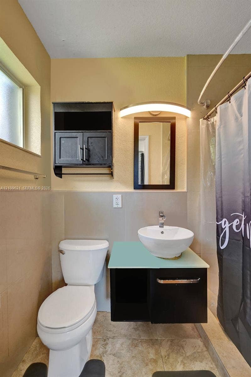 231 SW 64th Way, Pembroke Pines, FL 33023 Wall-mounted floating vanity with a frosted glass countertop and white vessel sink, complemented by matching dark cabinetry, a wall-mounted arched light fixture, and stone-finish floor tiling