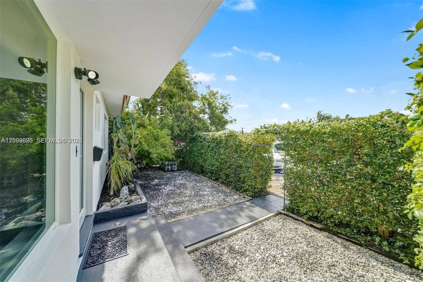 Covered patio area featuring stone ground cover, mature privacy hedging, and exterior wall-mounted lighting