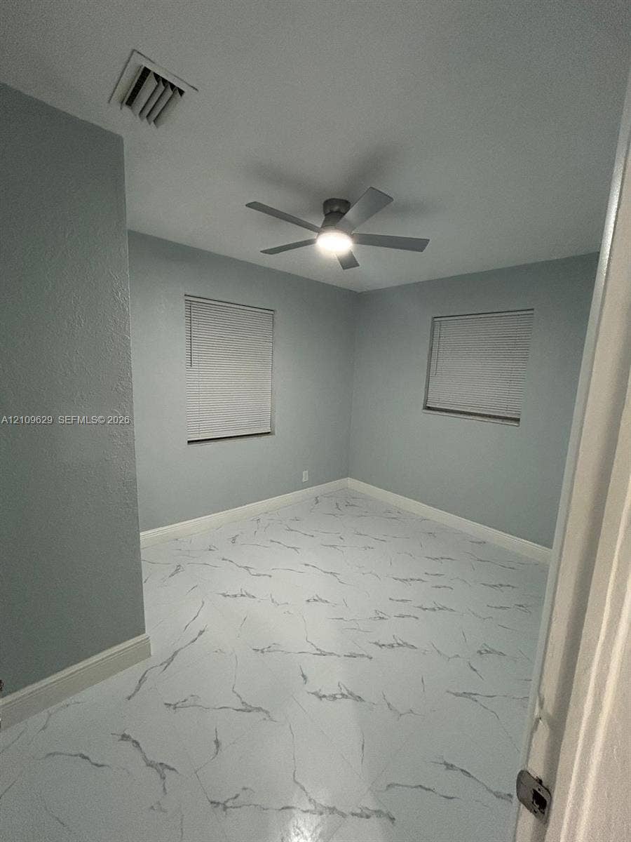 1741 NW 24th Terrace, Fort Lauderdale, FL 33311 Neutral-toned room featuring large-format tile flooring with marble-patterned veining, dual windows with fitted blinds, and a multi-blade ceiling fan with integrated lighting