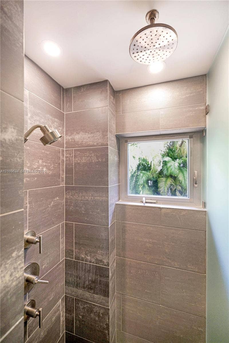 Walk-in shower featuring large-format ceramic tile, ceiling-mounted rainfall showerhead, wall-mounted handheld sprayer, multi-handle temperature control system, and a fixed window