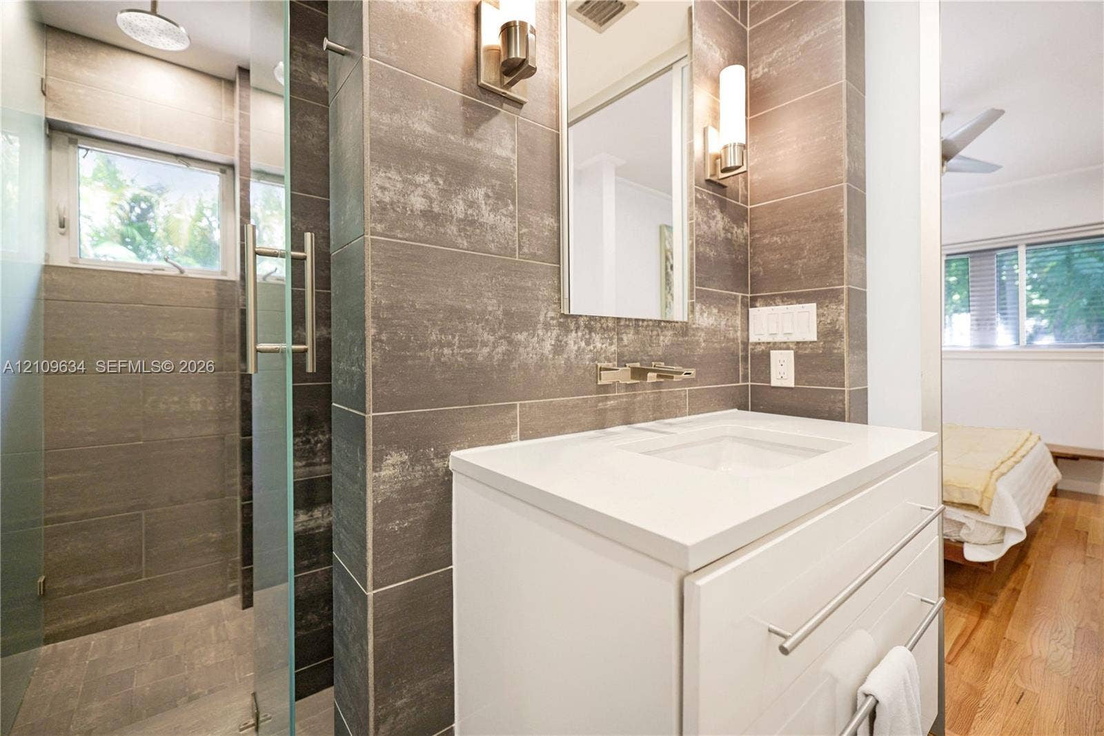 Modern vanity with a rectangular sink, wall-mounted faucet, and quartz countertop featuring full-height large-format wall tiling and a glass-enclosed walk-in shower with a rainfall showerhead