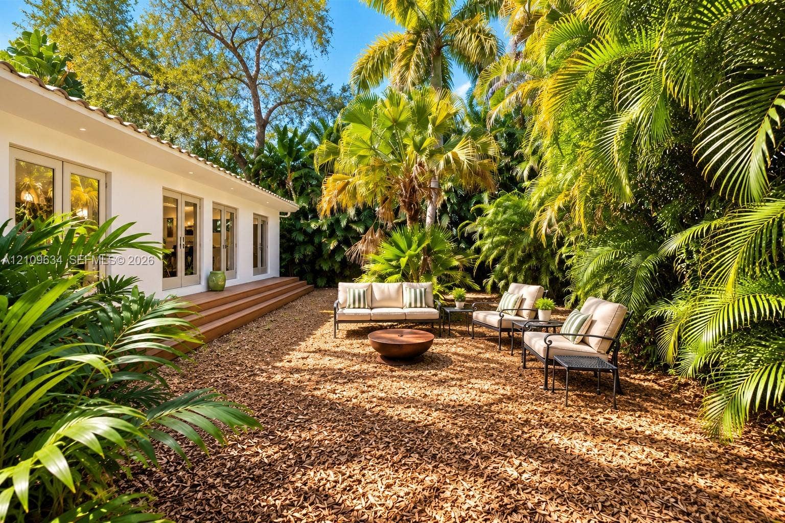 Exterior patio area featuring a low-profile wood-finish deck, multi-panel glass French doors, and mature tropical landscaping