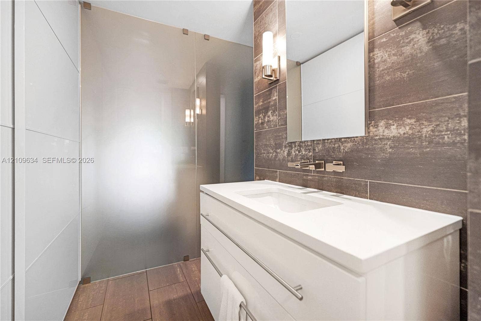 Modern vanity featuring a white finish and integrated sink, paired with a frameless glass shower enclosure, metallic wall-mounted faucet, rectangular wall mirror, and large-format tile wall cladding