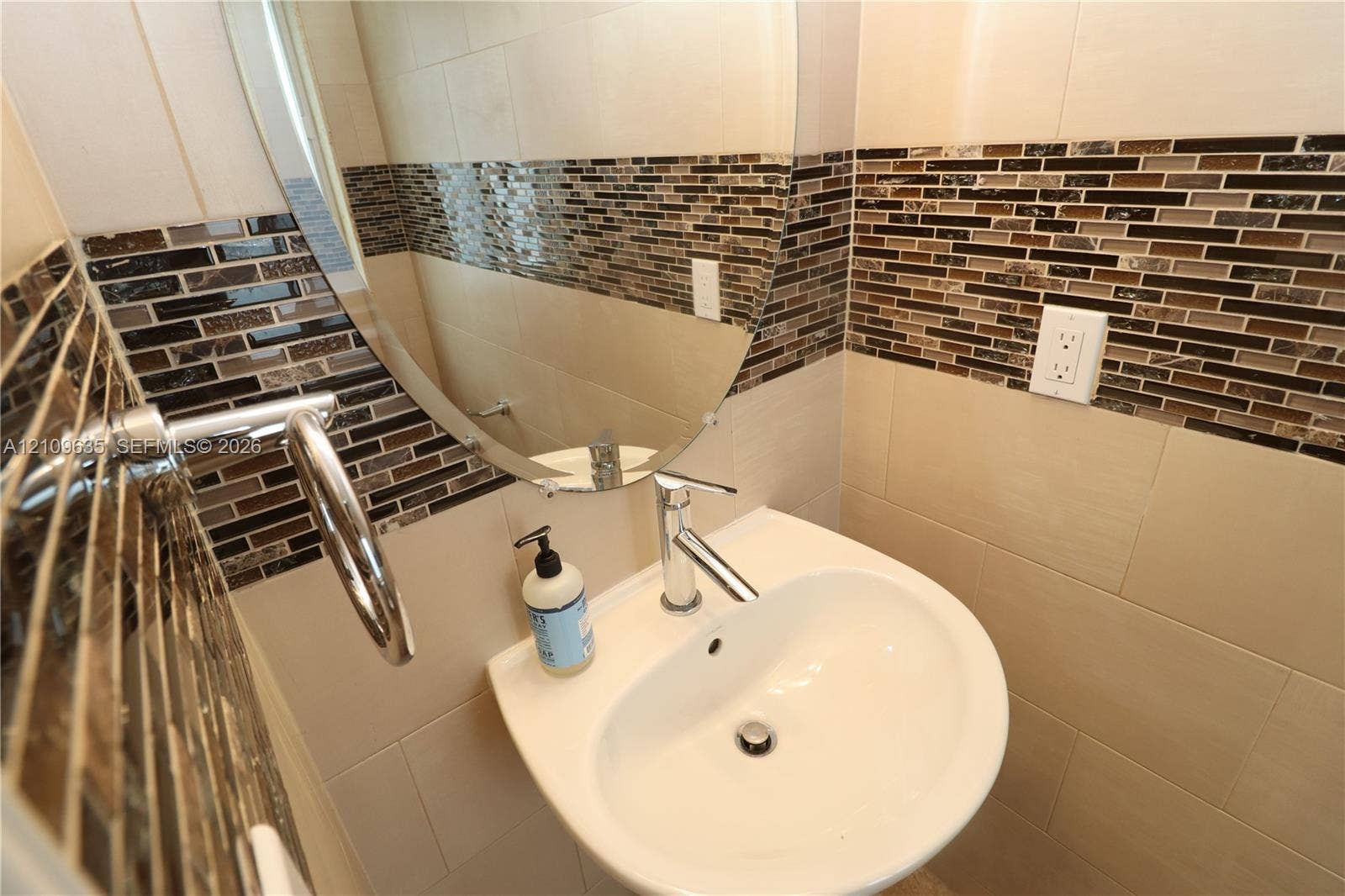 611 NE 28th Street, #1, Wilton Manors, Wilton Manors, FL 33334 Wall-mounted white basin sink featuring a single-handle chrome faucet, oval frameless mirror, and horizontal mosaic tile backsplash with large-format neutral wall tiles