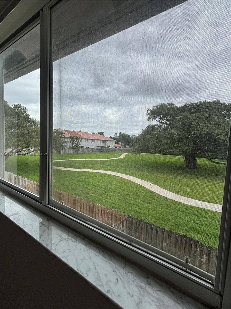 11467 NW 51st Terrace, Doral, Doral, FL 33178 Horizontal sliding window featuring a stone-finish sill, revealing a manicured lawn with a curved paved path, mature canopy tree, and wood fencing