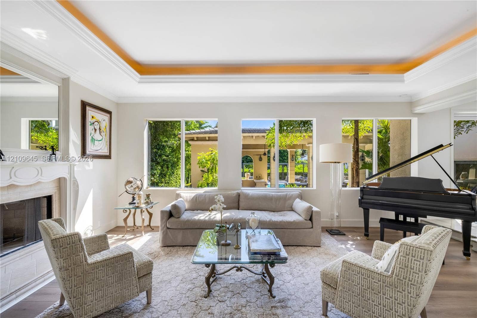 332 Costanera Road, Coral Gables, FL 33143 Living area featuring tray ceilings, wood-finish flooring, and a fireplace with a detailed mantel, illuminated by large windows