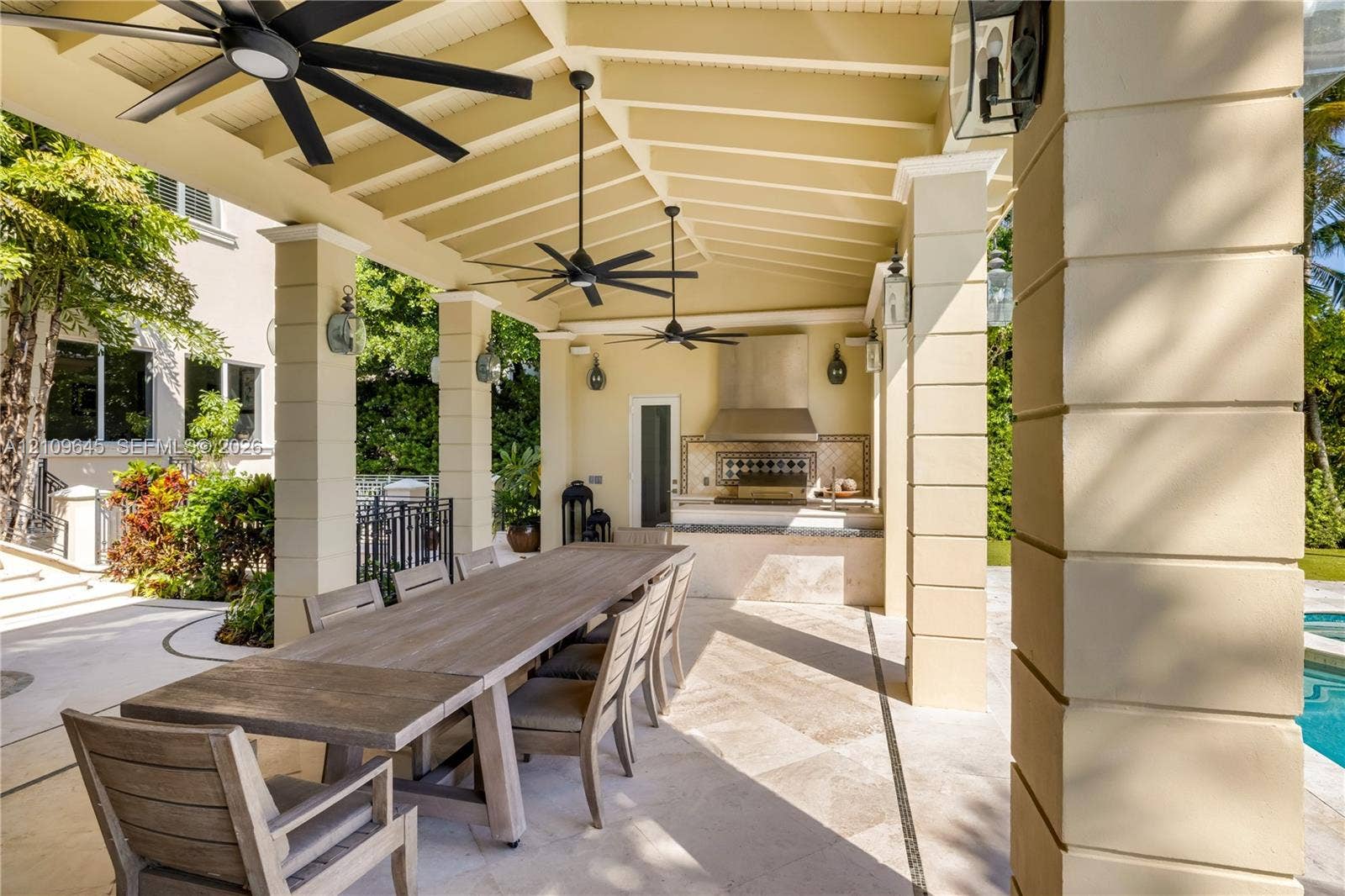 332 Costanera Road, Coral Gables, FL 33143 Covered outdoor kitchen and dining area featuring a stone-tile patio, built-in stainless steel range hood, stone-clad counter, multi-blade ceiling fans, and decorative wall-mounted lighting