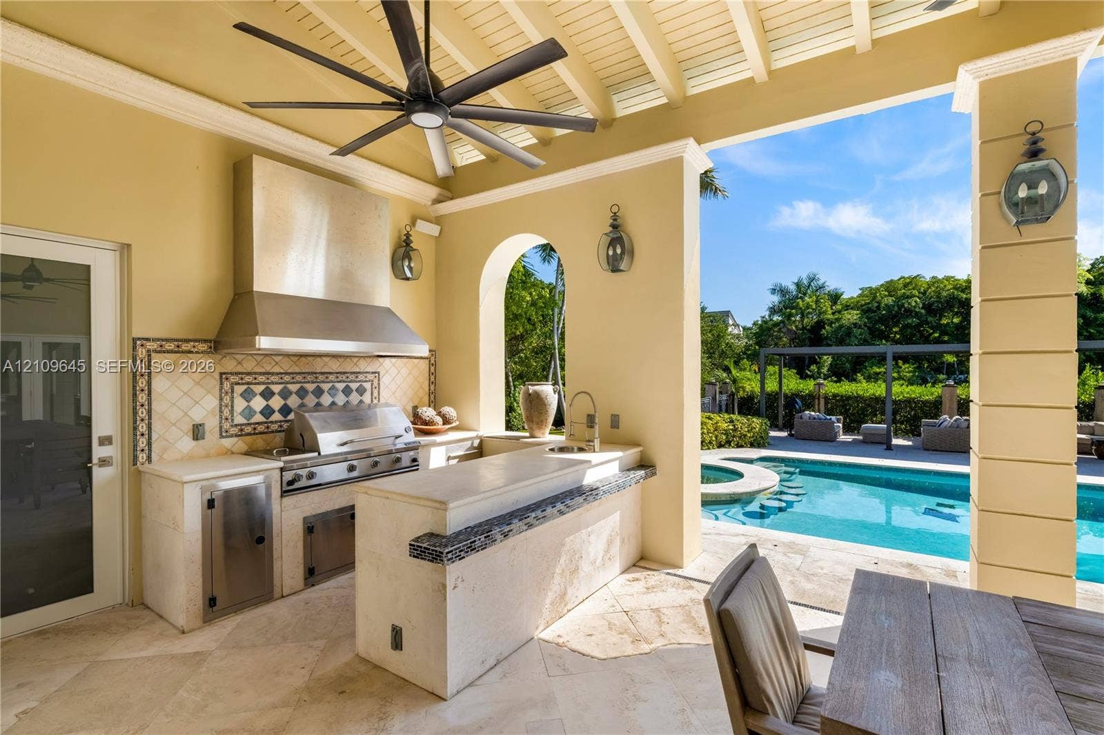 332 Costanera Road, Coral Gables, FL 33143 Covered outdoor kitchen featuring stone countertops, a stainless steel grill, a tile backsplash, and a built-in sink, accented by an arched opening and a pool with integrated seating