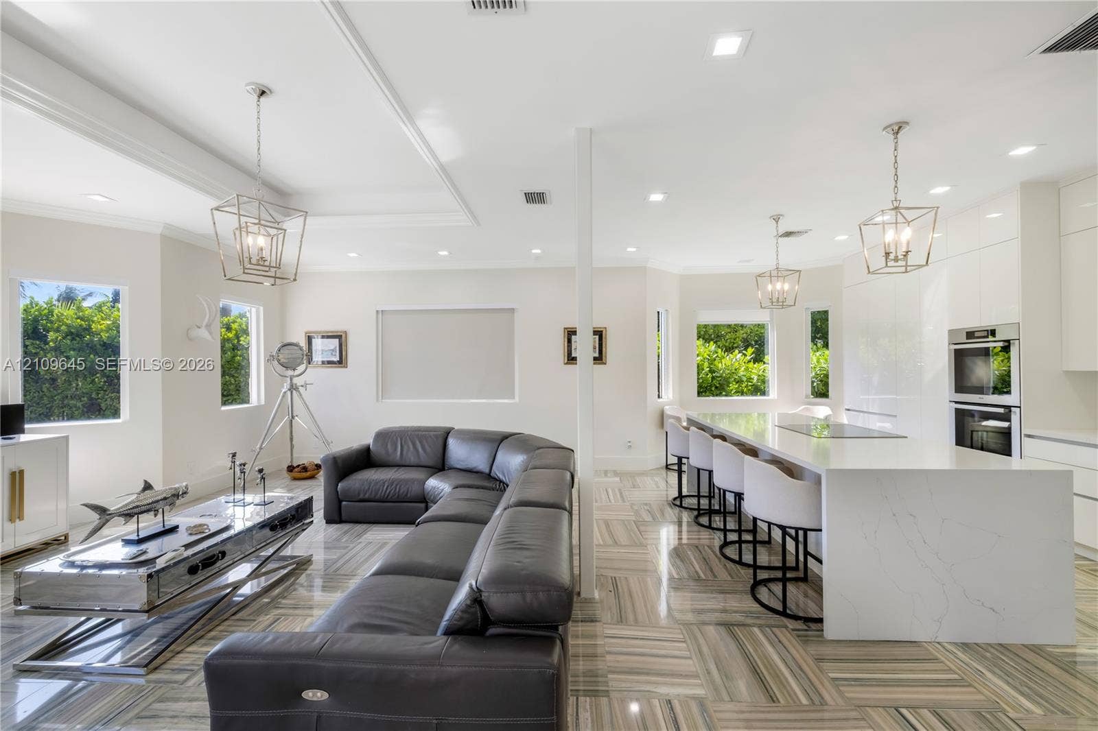 332 Costanera Road, Coral Gables, FL 33143 Open-concept interior featuring large-format tile flooring, a waterfall-edge stone island with integrated cooktop, dual built-in wall ovens, recessed lighting, and geometric metal pendant fixtures