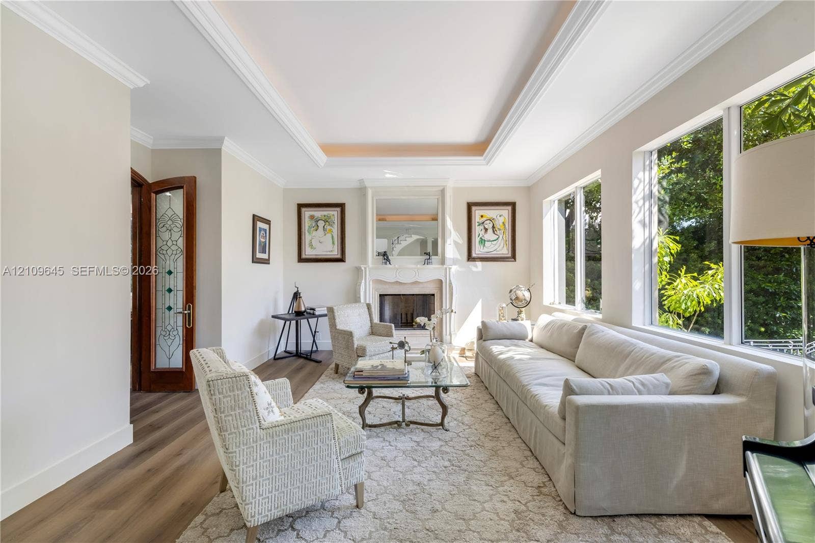 332 Costanera Road, Coral Gables, FL 33143 Living area featuring wood-finish flooring, a tray ceiling with crown molding, a wood-burning fireplace with a mantel, and oversized windows