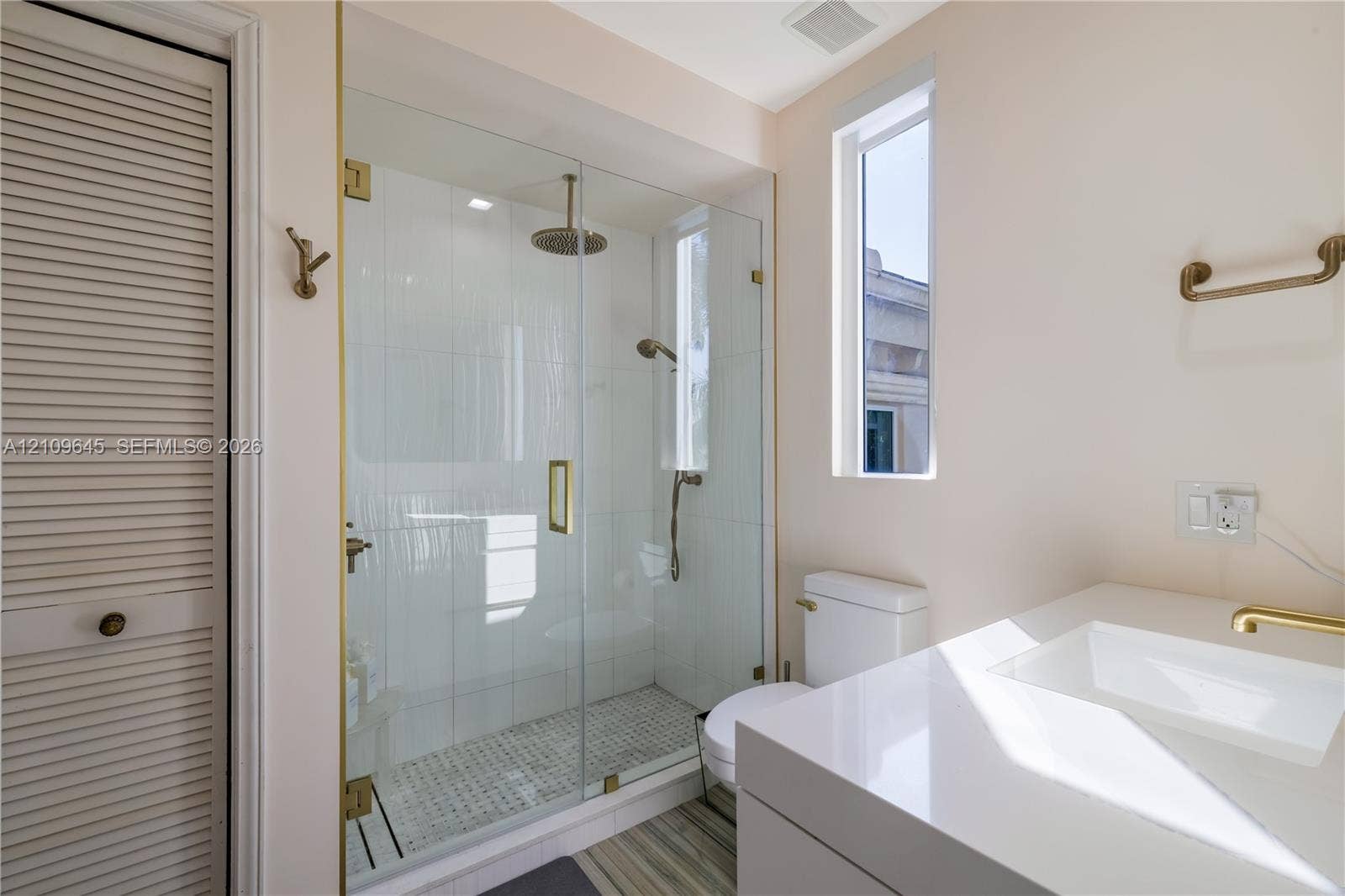 332 Costanera Road, Coral Gables, FL 33143 Walk-in glass shower featuring a rain-style showerhead and handheld sprayer with brass hardware, complemented by a white vanity with a rectangular integrated sink and a louvered-door closet