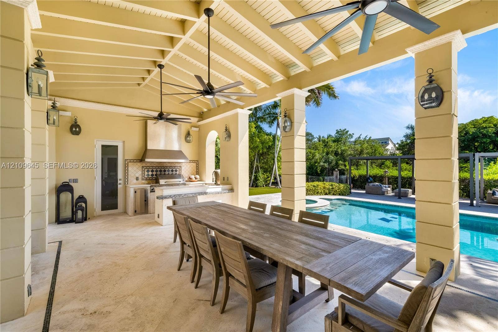 332 Costanera Road, Coral Gables, FL 33143 Covered outdoor patio featuring stone flooring, vaulted beamed ceiling with multiple ceiling fans, built-in kitchen with stainless steel range hood, and tile backsplash