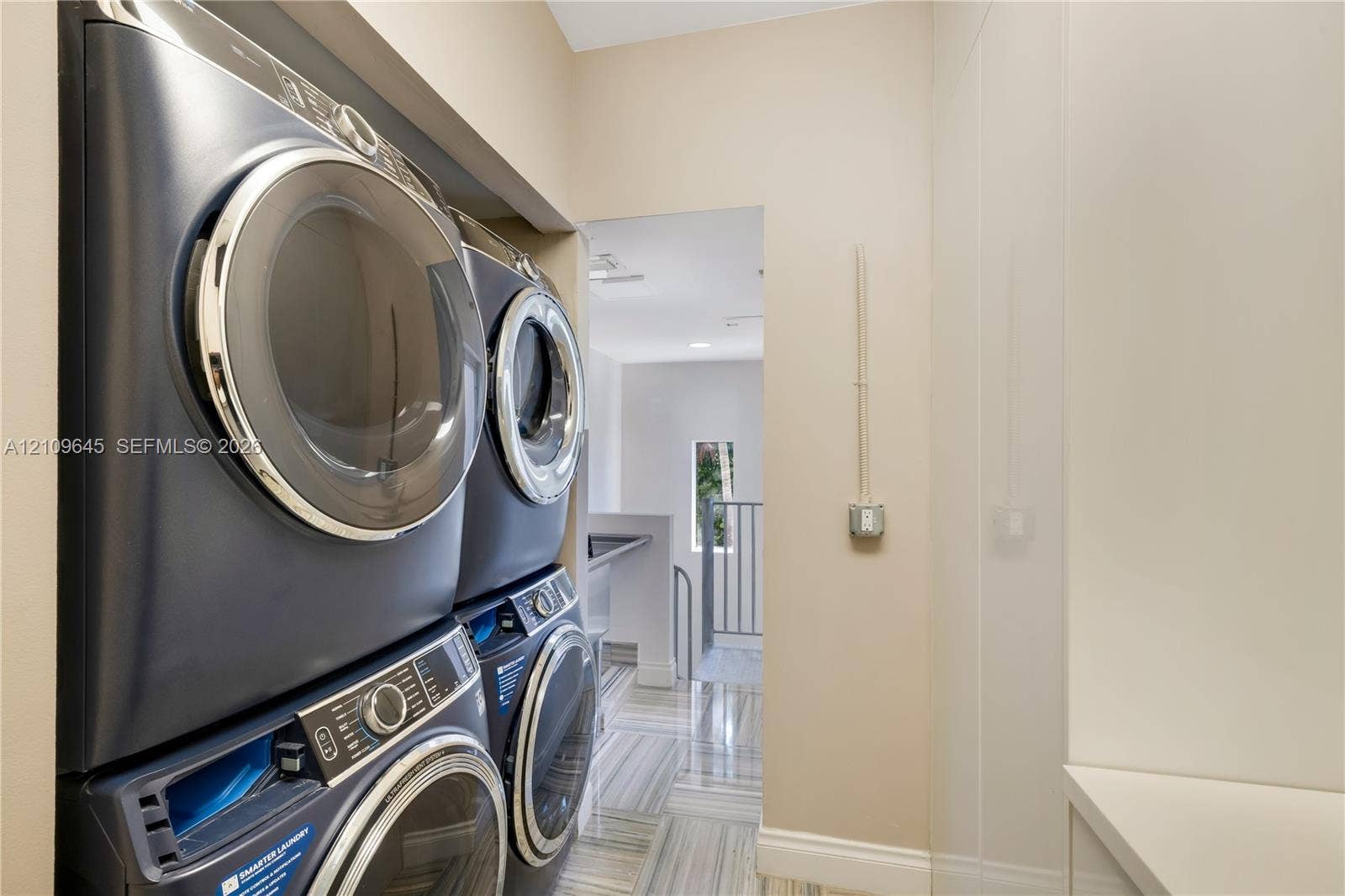 332 Costanera Road, Coral Gables, FL 33143 Laundry area featuring stacked metallic-finish washing machine and dryer, floor-to-ceiling built-in cabinetry, and neutral stone-patterned tile flooring
