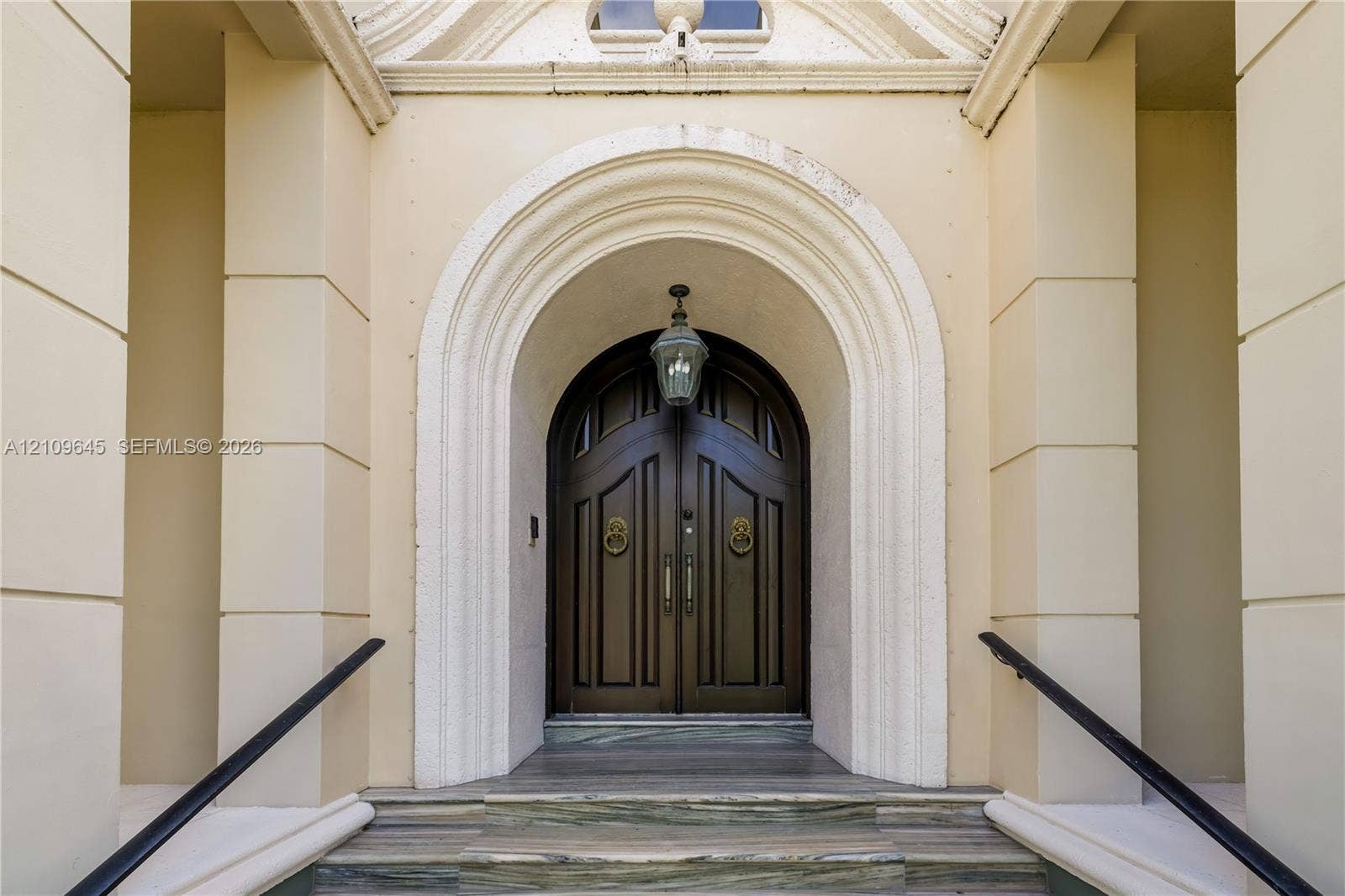332 Costanera Road, Coral Gables, FL 33143 Double arched solid wood doors featuring brass lion-head knockers, set within a recessed masonry entryway with decorative molding, stone-stepped risers, and mounted pendant lighting