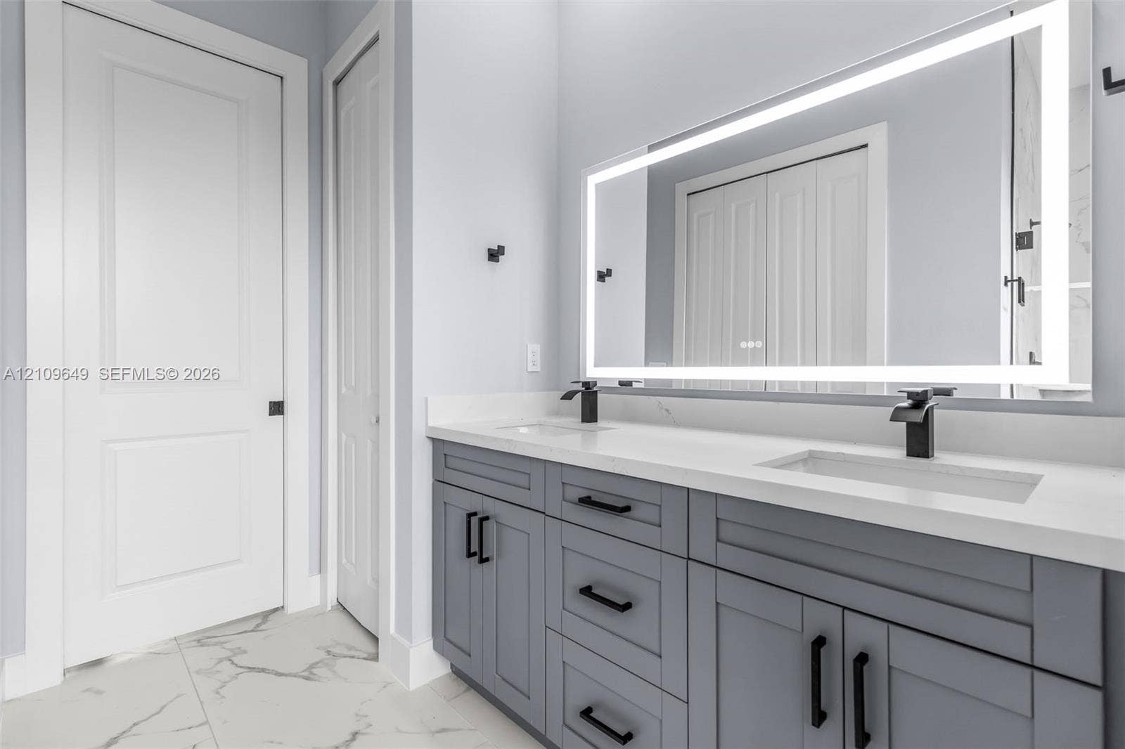 2818 42nd St Sw Lehigh Acres, Lehigh Acres, FL 33976 Double vanity with grey shaker-style cabinetry, white solid-surface countertop, integrated dual sinks, matte black fixtures, and a large backlit rectangular mirror