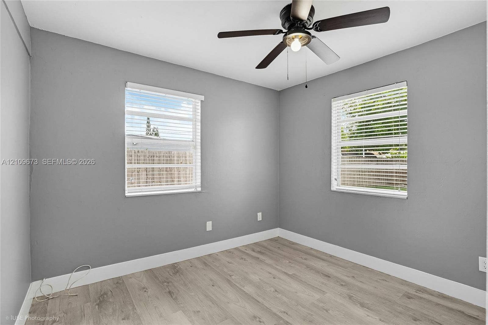 1748 NE 49th Street, Pompano Beach, FL 33064 Interior space featuring wood-finish flooring, neutral gray wall paint, dual windows with horizontal blinds, and a central ceiling fan with an integrated light fixture
