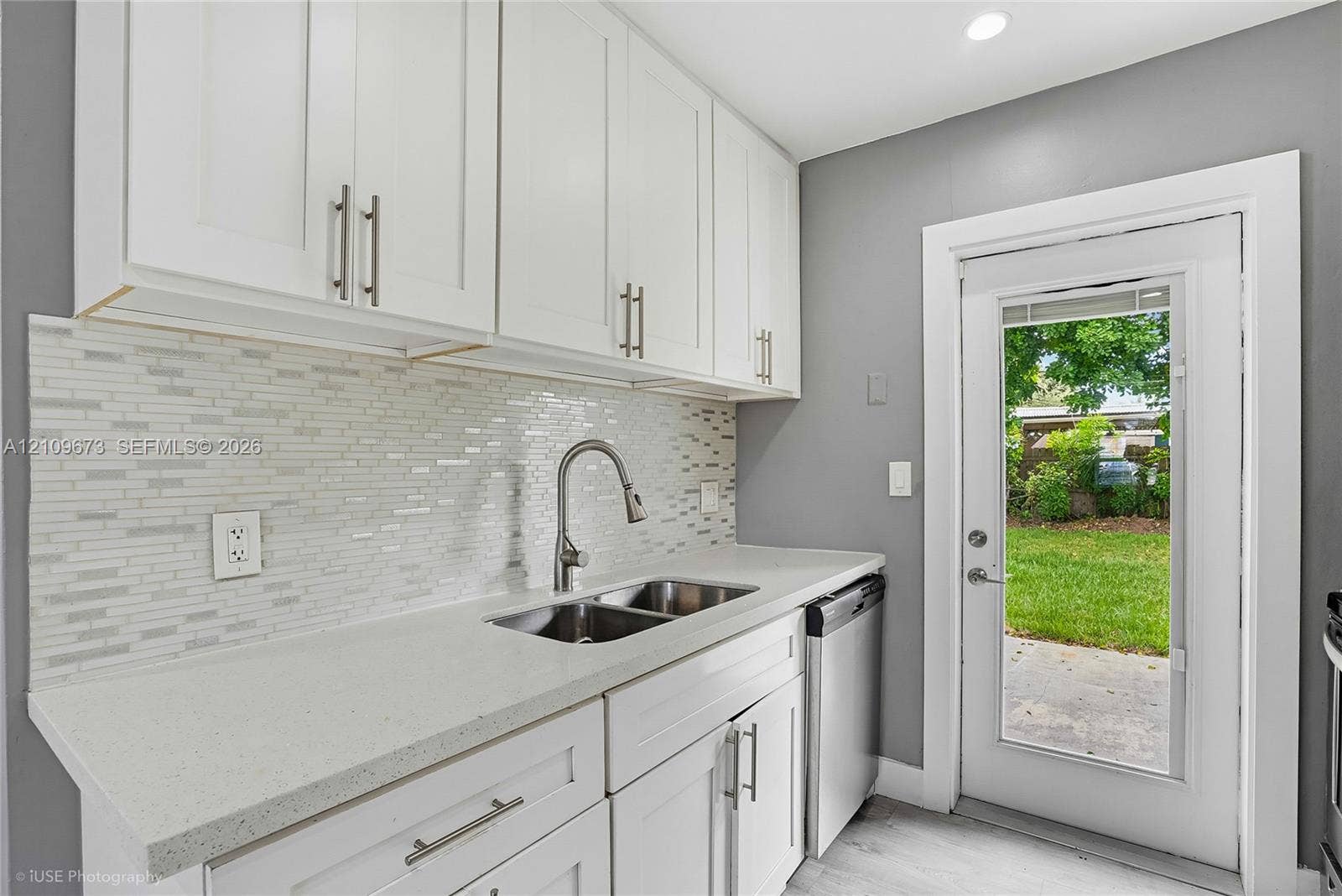 1748 NE 49th Street, Pompano Beach, FL 33064 Kitchen featuring white shaker-style cabinetry, solid-surface countertops, horizontal glass tile backsplash, dual-basin stainless steel sink with high-arc faucet, and an exterior glass-panel door