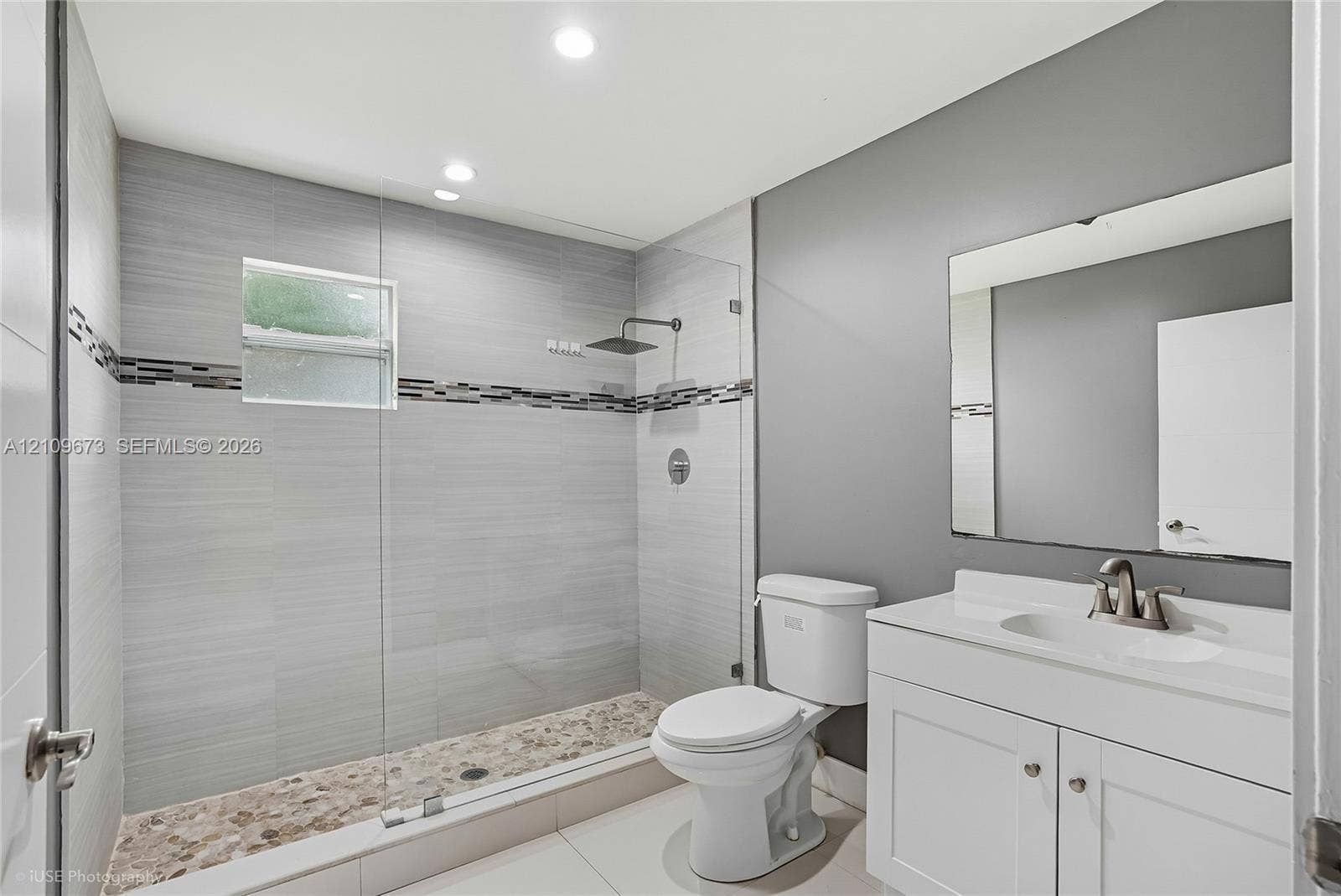 1748 NE 49th Street, Pompano Beach, FL 33064 Walk-in shower with floor-to-ceiling gray tile, pebble-stone flooring, frameless glass enclosure, and a rain-style showerhead, complemented by a white vanity with an integrated sink and rectangular wall mirror