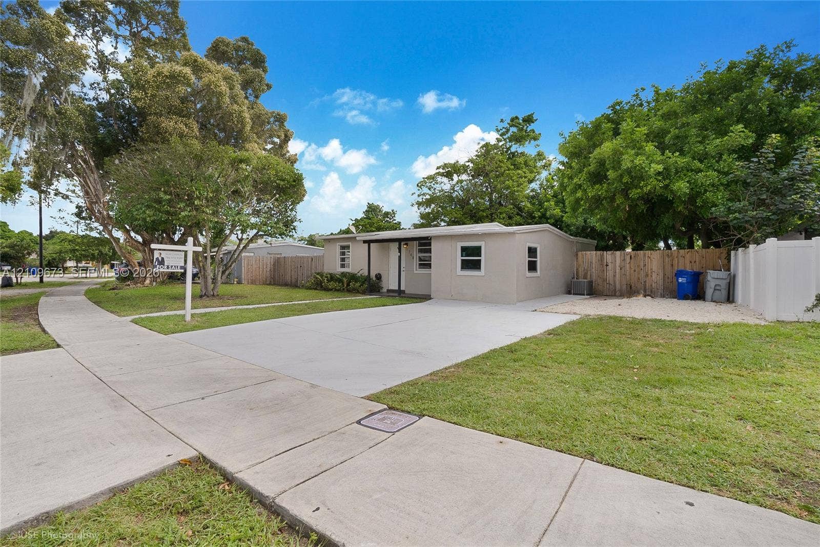 1748 NE 49th Street, Pompano Beach, FL 33064 Single-story exterior featuring a concrete driveway, lawn, mature trees, and wood-panel perimeter fencing
