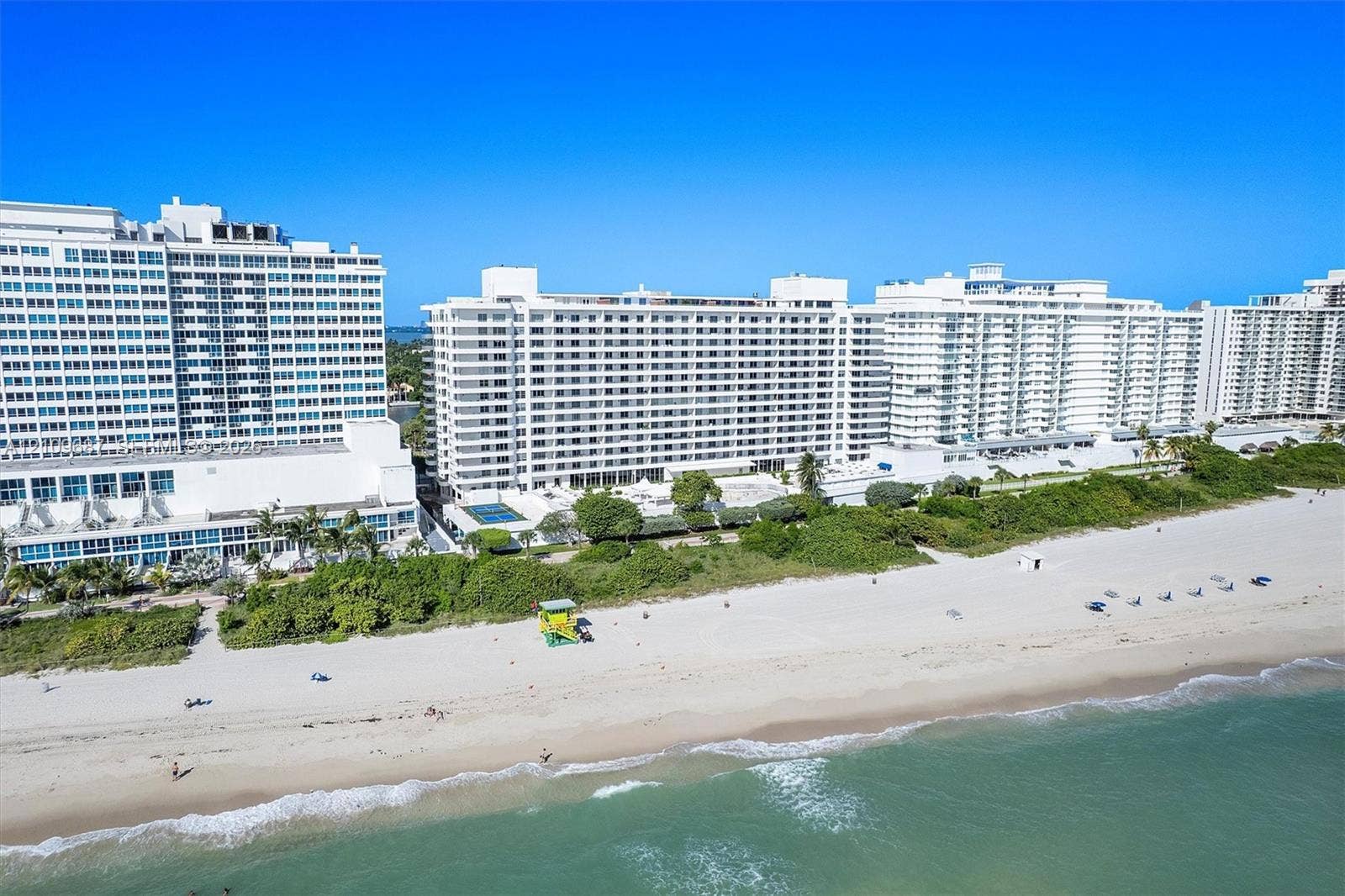 5555 Collins Avenue, #5C, Miami Beach, FL 33140 Multi-story beachfront residential buildings featuring white stucco facades, balconies, dense coastal landscaping, and direct access to a sandy shoreline