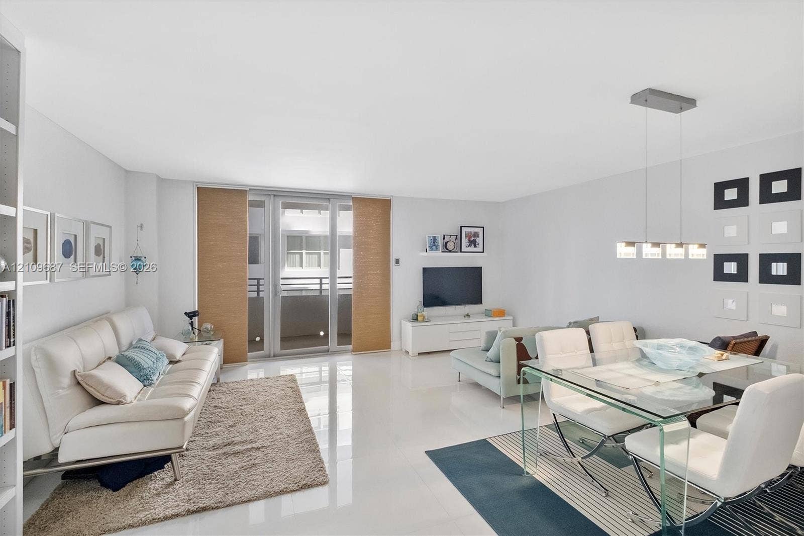 5555 Collins Avenue, #5C, Miami Beach, FL 33140 Open-concept living space featuring high-gloss tile flooring, floor-to-ceiling glass sliding doors, integrated wall shelving, and a modern multi-light linear pendant fixture
