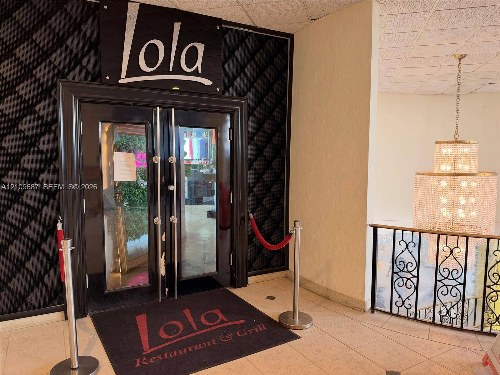 5555 Collins Avenue, #5C, Miami Beach, FL 33140 Double-door glass entrance with black finish framing, tufted black wall paneling, tile flooring, and decorative metal stair railing with integrated multi-tiered crystal chandelier