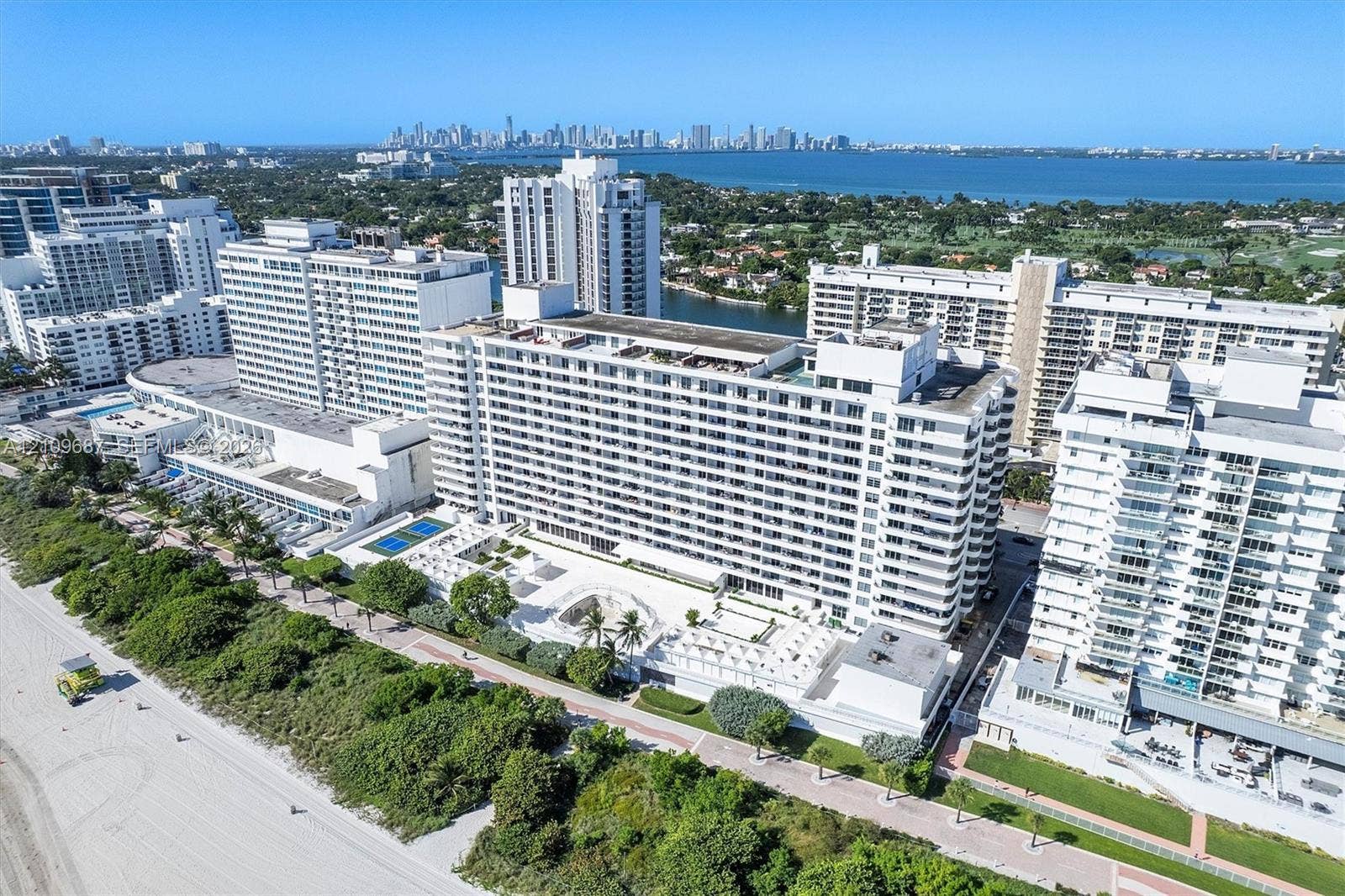 5555 Collins Avenue, #5C, Miami Beach, FL 33140 Aerial perspective of coastal high-rise building featuring direct beach access, outdoor swimming pools, multi-purpose sport courts, and expansive shoreline scenery