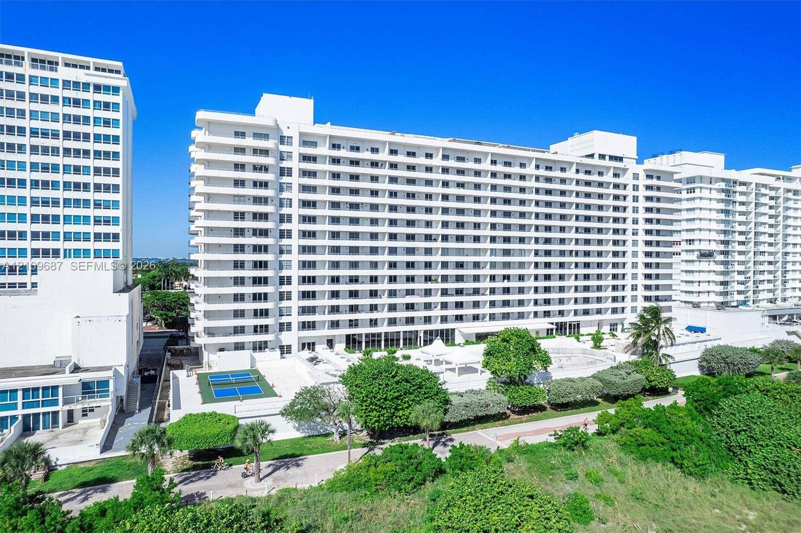 5555 Collins Avenue, #5C, Miami Beach, FL 33140 High-rise residential exterior featuring expansive tiered balconies, ground-level tennis courts, manicured landscaping, and mature palm trees