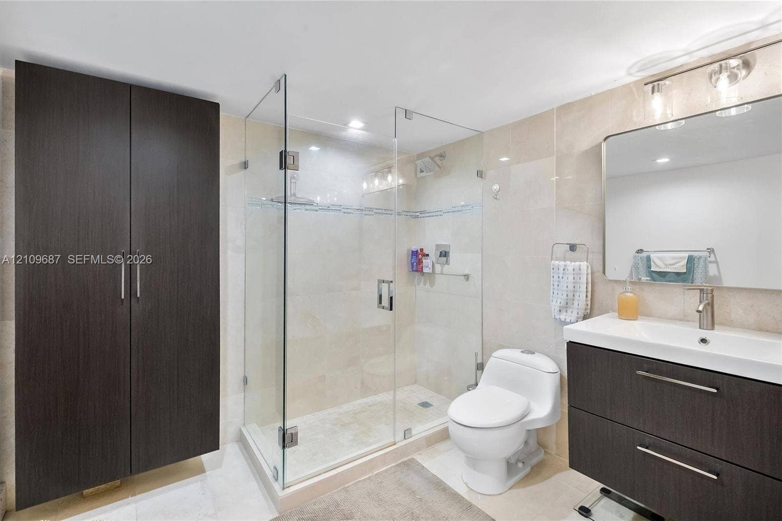 5555 Collins Avenue, #5C, Miami Beach, FL 33140 Bathroom featuring a glass-enclosed shower, dark wood-finish cabinetry, a rectangular vanity sink with a metallic faucet, and stone-tile walls