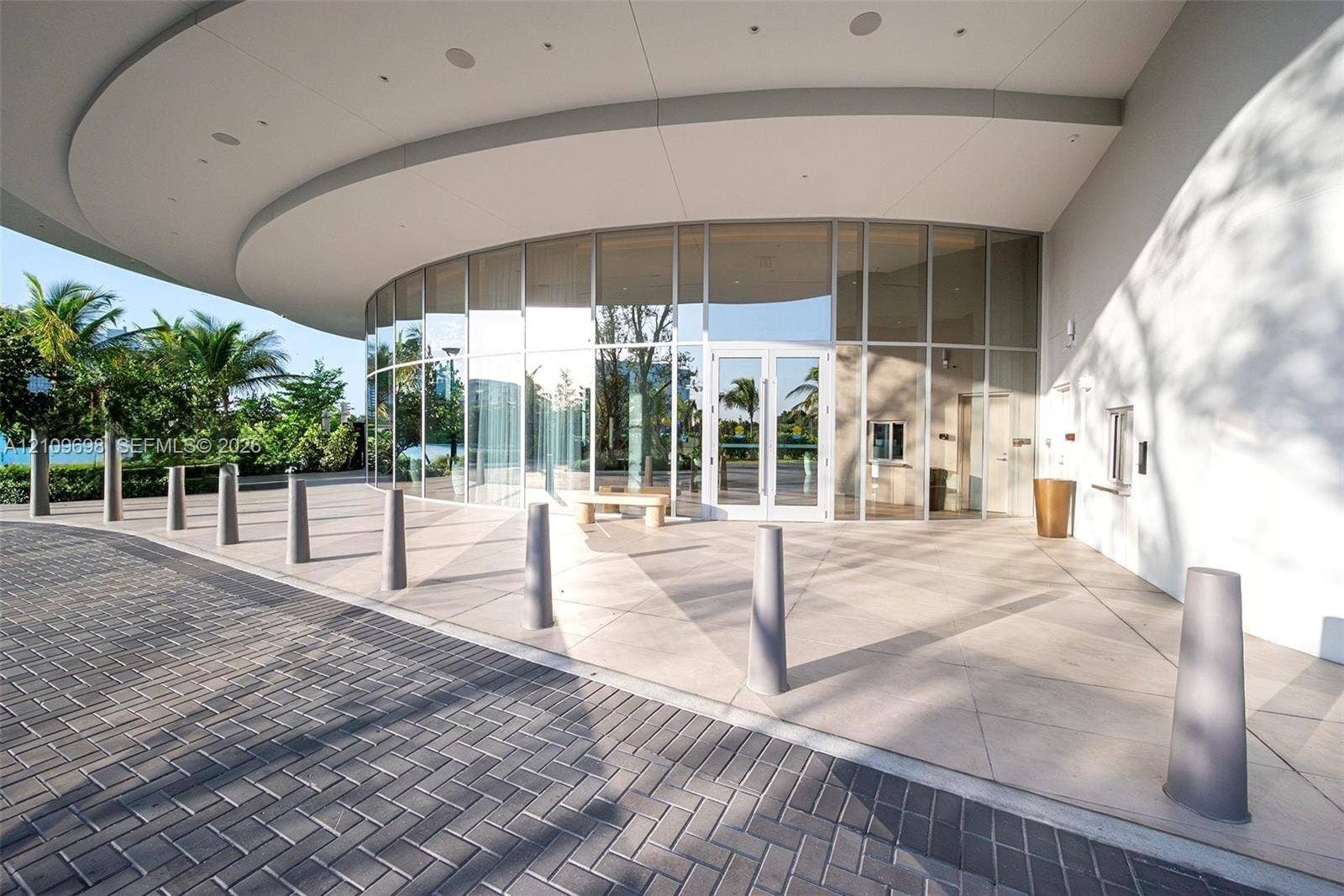 2411 Laguna Circle, #810, North Miami, FL 33181 Curved glass faade featuring floor-to-ceiling windows and a deep structural overhang, complemented by a herringbone-patterned paver driveway and a wide concrete pedestrian plaza
