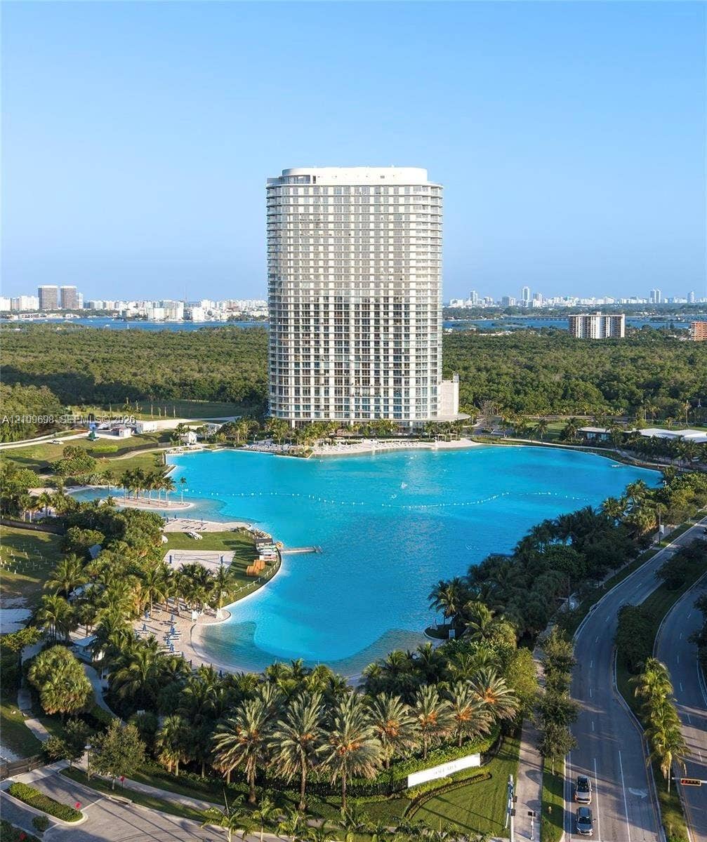 2411 Laguna Circle, #810, North Miami, FL 33181 High-rise residential tower featuring floor-to-ceiling windows, expansive man-made lagoon, white sand beaches, and lush tropical landscaping