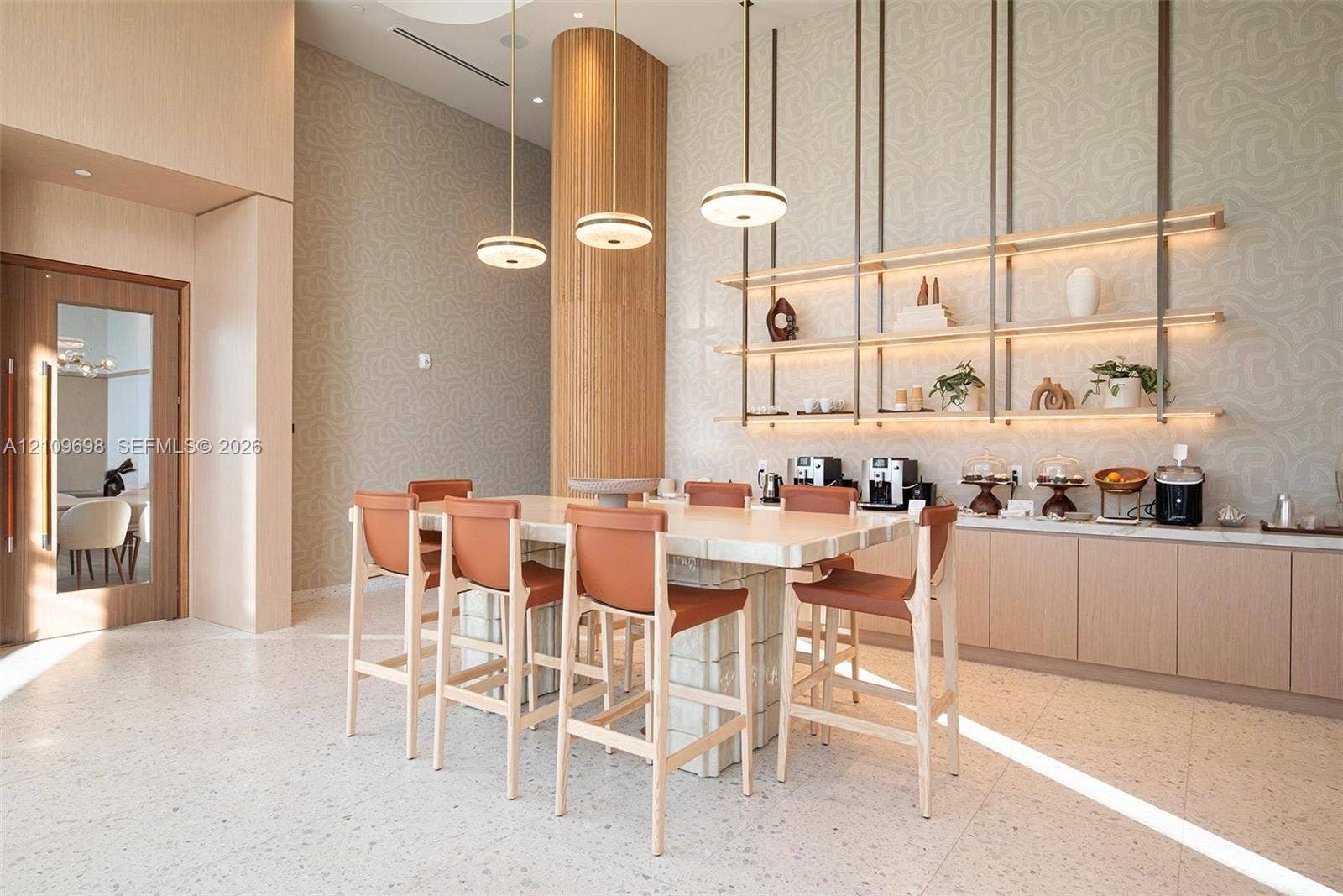 2411 Laguna Circle, #810, North Miami, FL 33181 High-ceiling common area featuring terrazzo-style flooring, a central island with a tiled base, integrated wall-mounted shelving with recessed lighting, and modern multi-tiered pendant fixtures